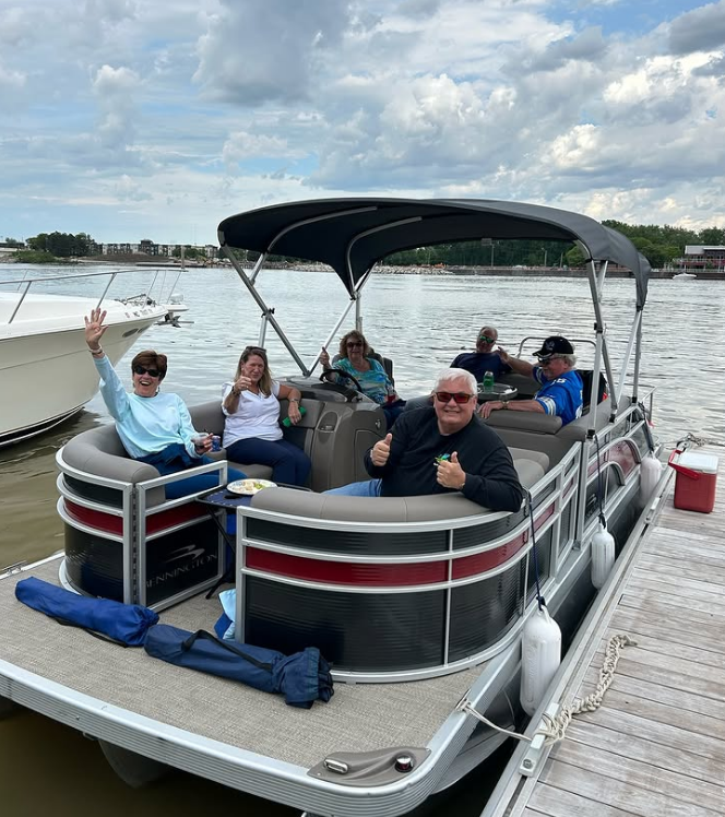 Group of six people on a pontoon boat anchored at a dock by a river, with a cloudy sky overhead.