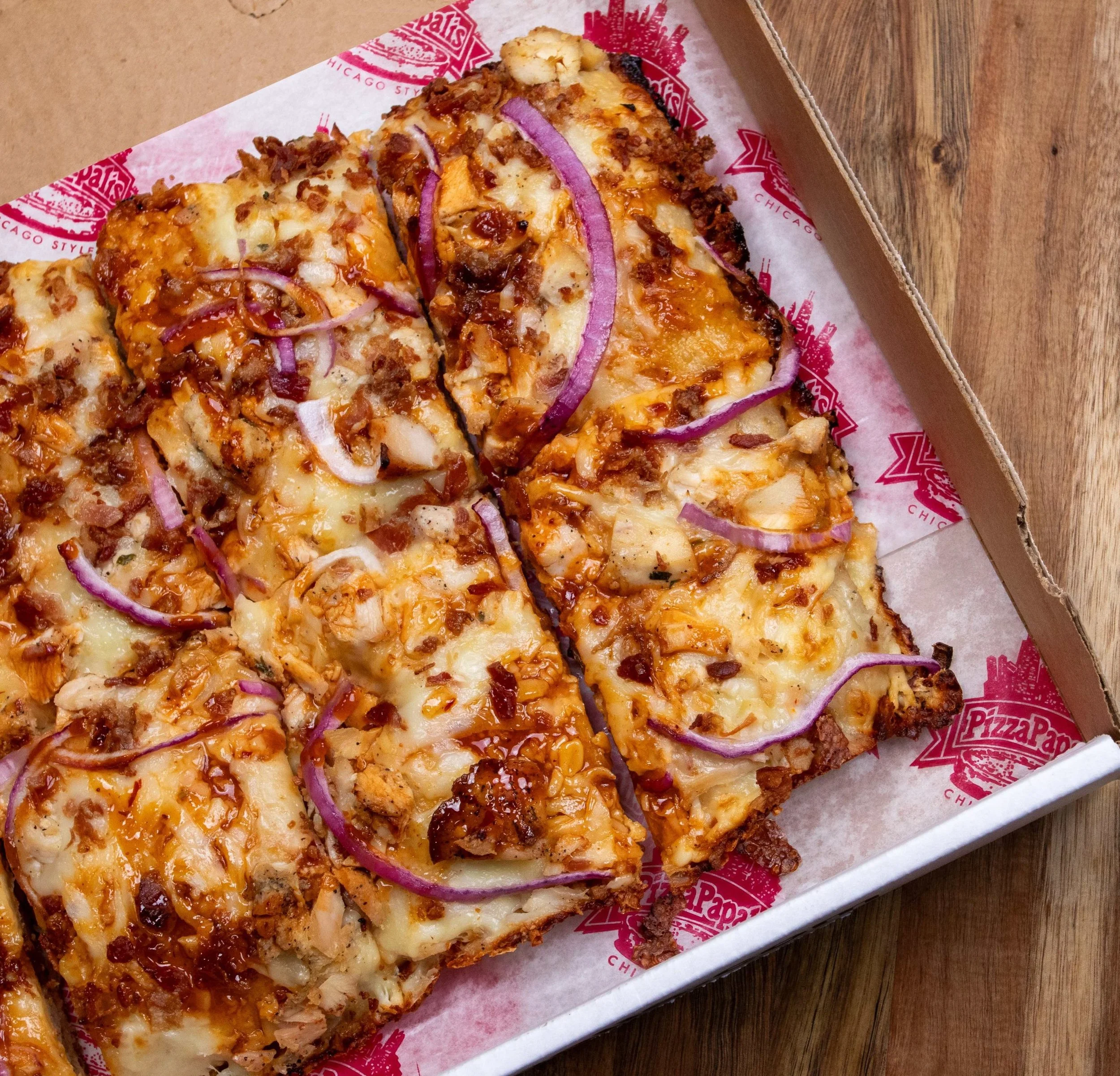 Close-up of a pizza with cheese, bacon, and red onions in a cardboard box on a wooden surface.