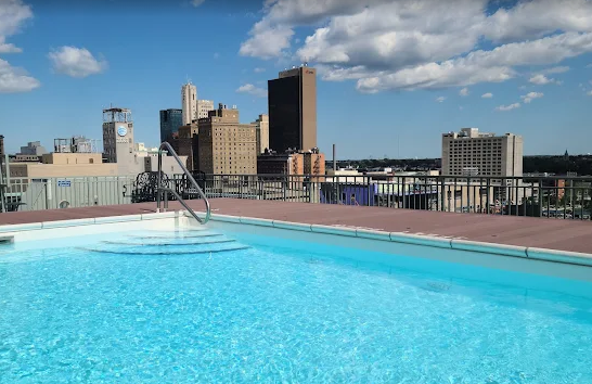 Swimming pool on a rooftop with a city skyline in the background under a partly cloudy sky.