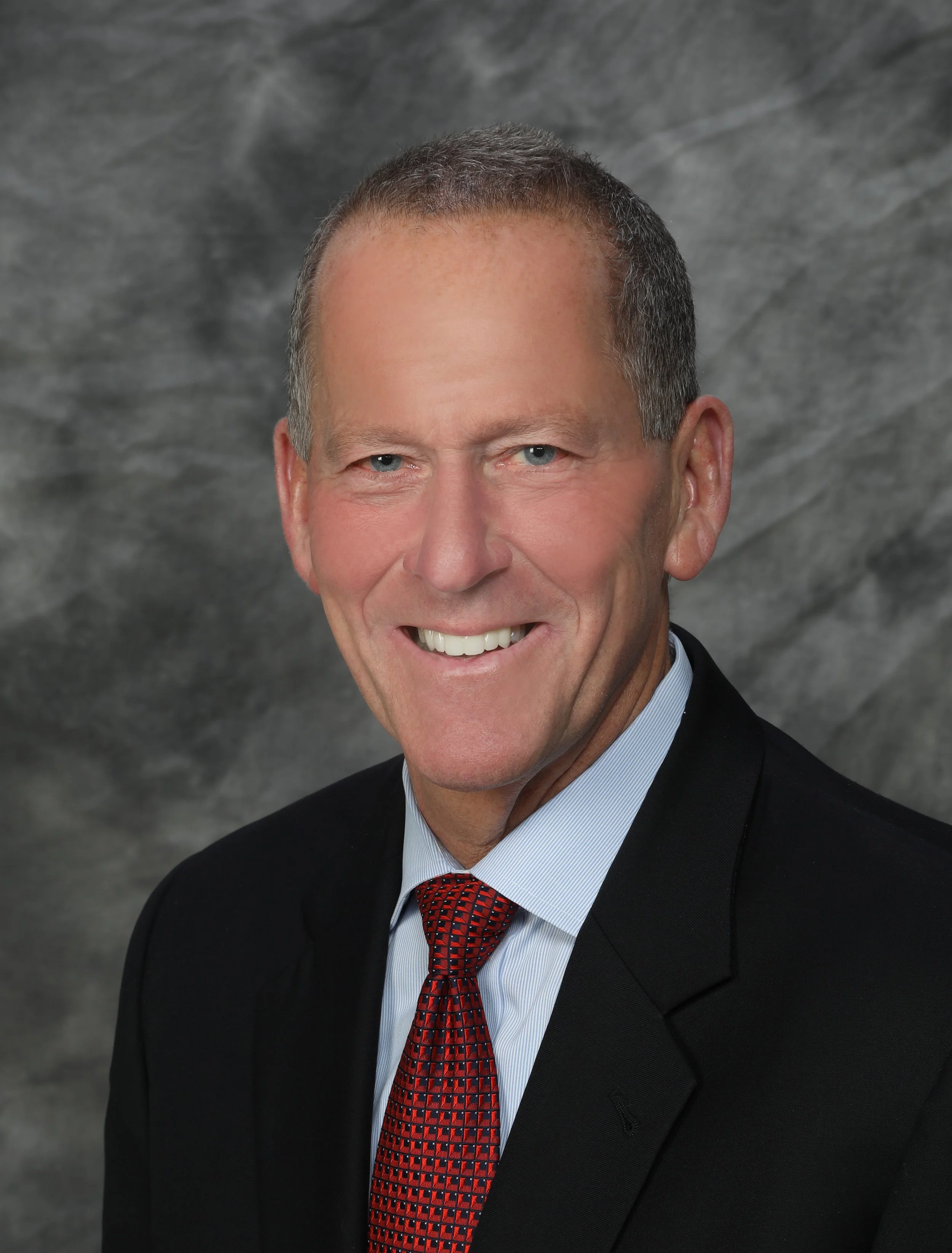 Professional headshot of a middle-aged man with short, light-colored hair, wearing a dark suit, white shirt, and patterned tie, smiling against a light gray background.