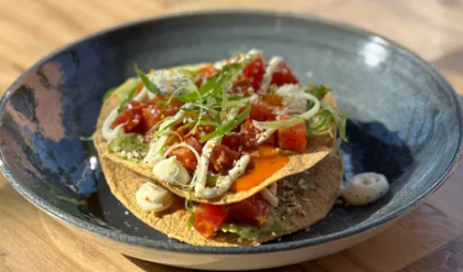 Open-faced taco on a blue plate with diced tomatoes, shredded cheese, green herbs, and sliced onions on a corn tortilla.