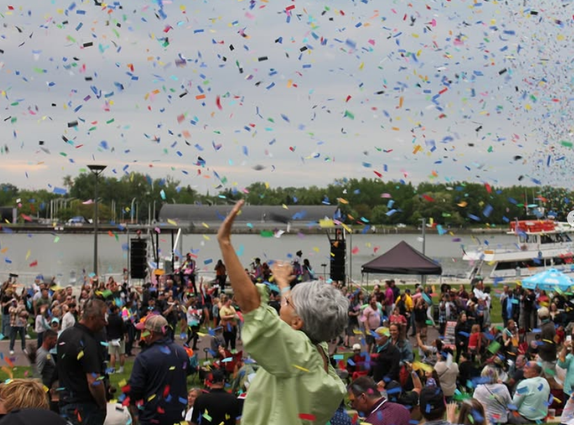 Crowd celebrating by a lake with colorful confetti falling from the sky, including an elderly woman raising her arm, tents, and boats in the background at a festival or event.