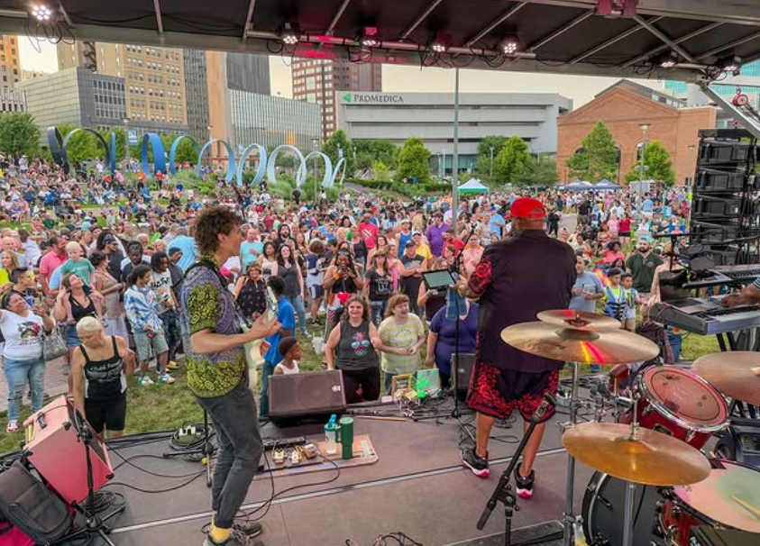 A musician with a red hat and patterned clothing performs on stage at an outdoor concert in a park, with a large crowd of diverse people enjoying the event, and city buildings in the background.