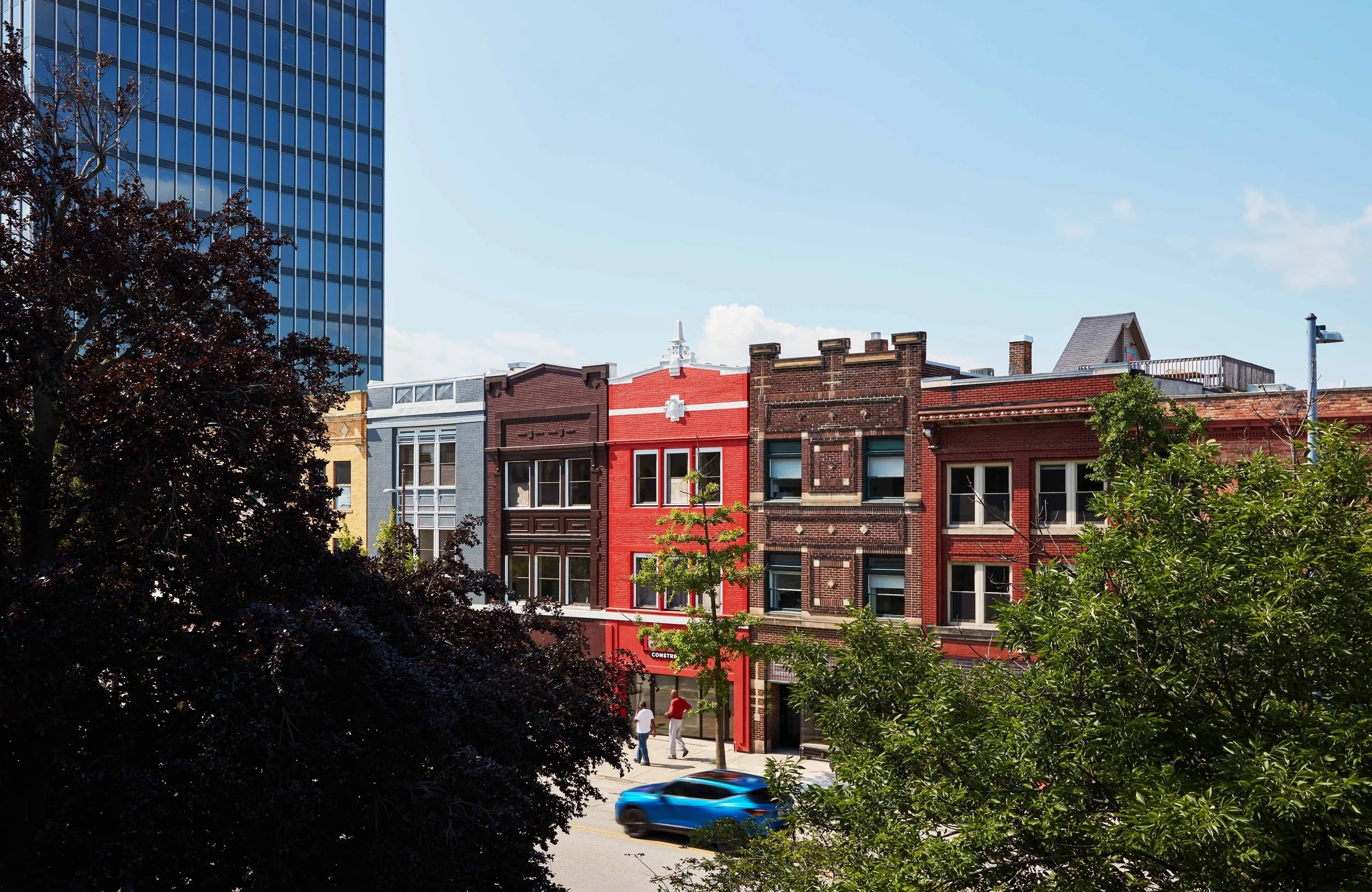 A street scene featuring a row of historic brick buildings, a red one in the center, with some trees and a blue car passing by in the foreground.