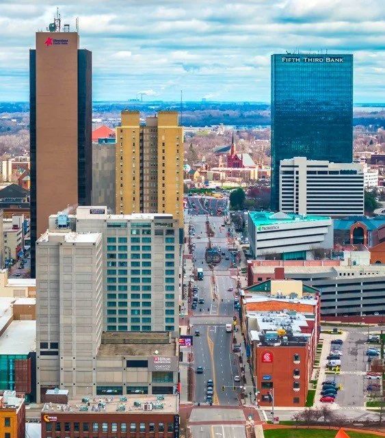 City skyline featuring tall buildings, including Fifth Third Bank and another large office building, with a busy street and traffic below, under a partly cloudy sky.