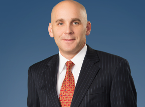 A middle-aged man wearing glasses, a dark suit, white shirt, and a red striped tie, standing against a dark blue background.