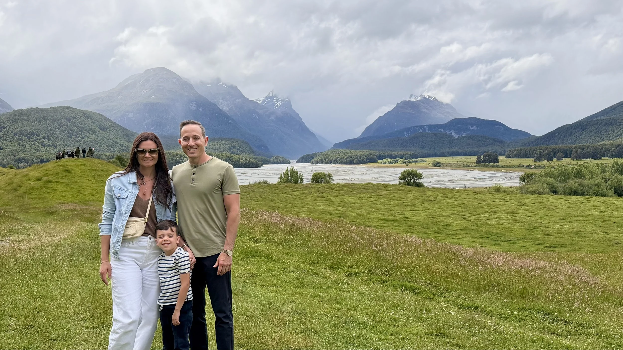 A family of three standing on a grassy hill with mountains, trees, and a river in the background, under cloudy skies.