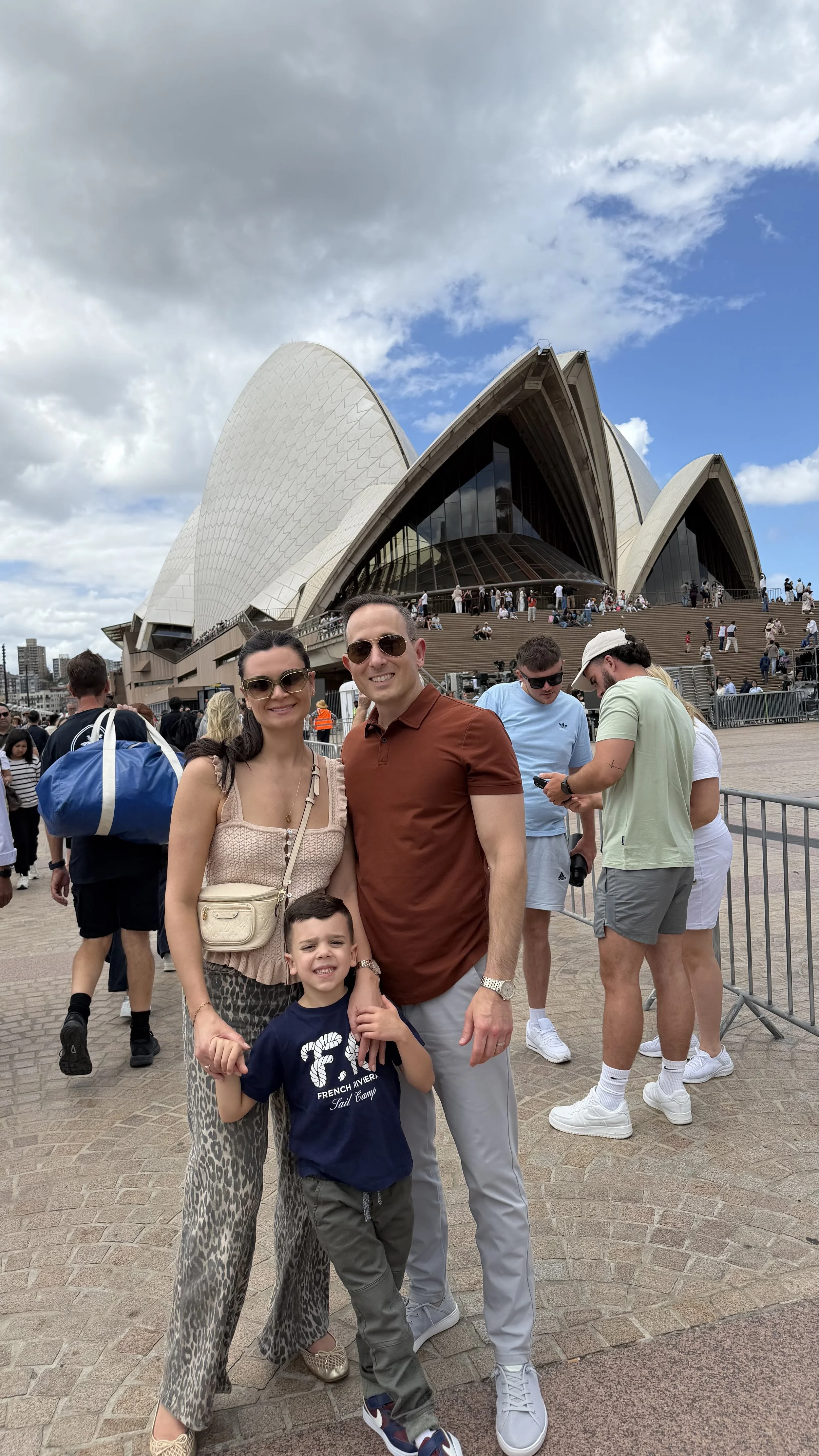 A family of three, a woman, a man, and a young boy, posing for a photo in front of the Sydney Opera House in Australia. There are other tourists and the iconic building in the background.