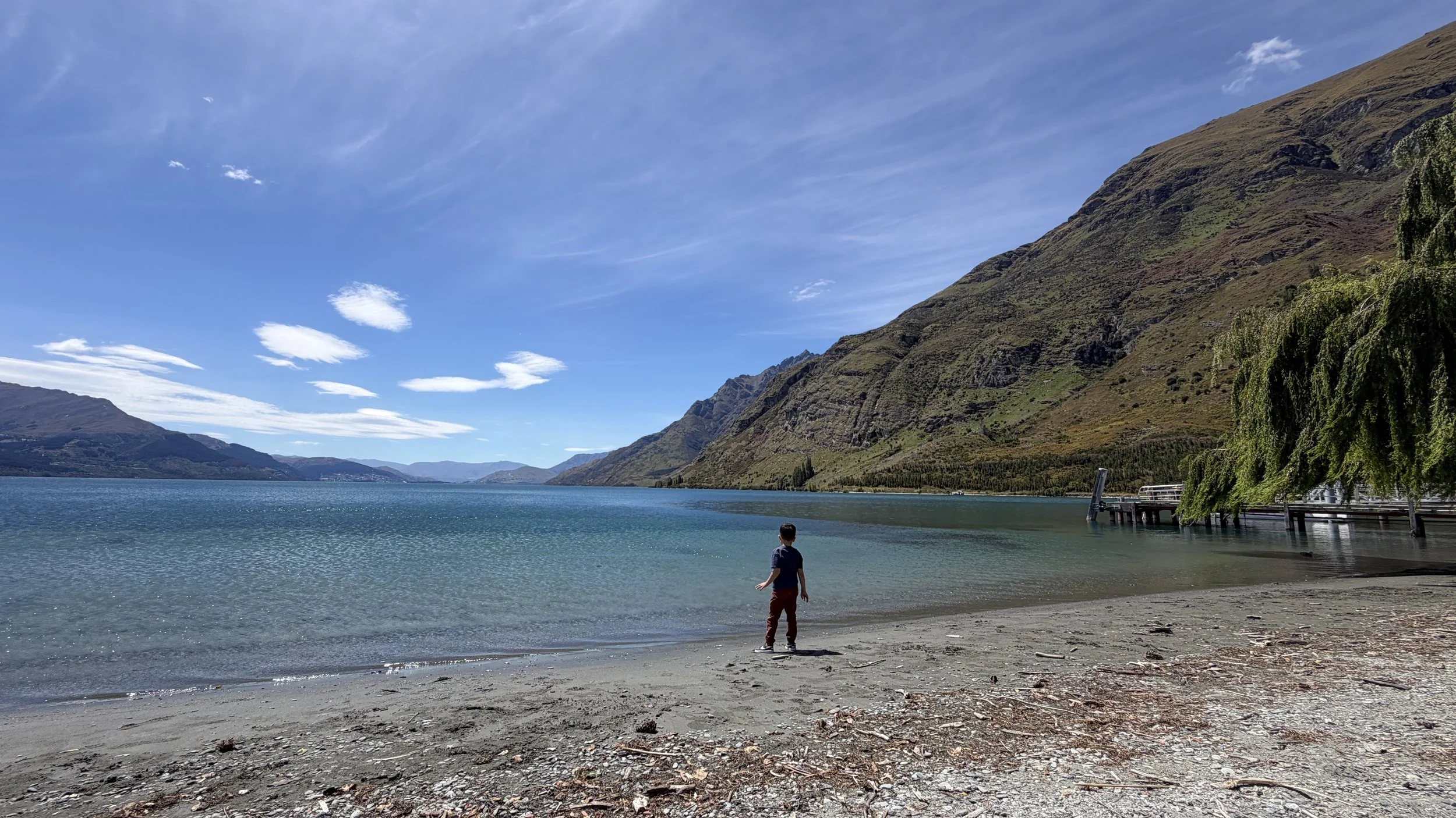 Child standing on a sandy beach by a large lake or river, with mountains and a partly cloudy sky in the background, and a pier extending into the water on the right side.