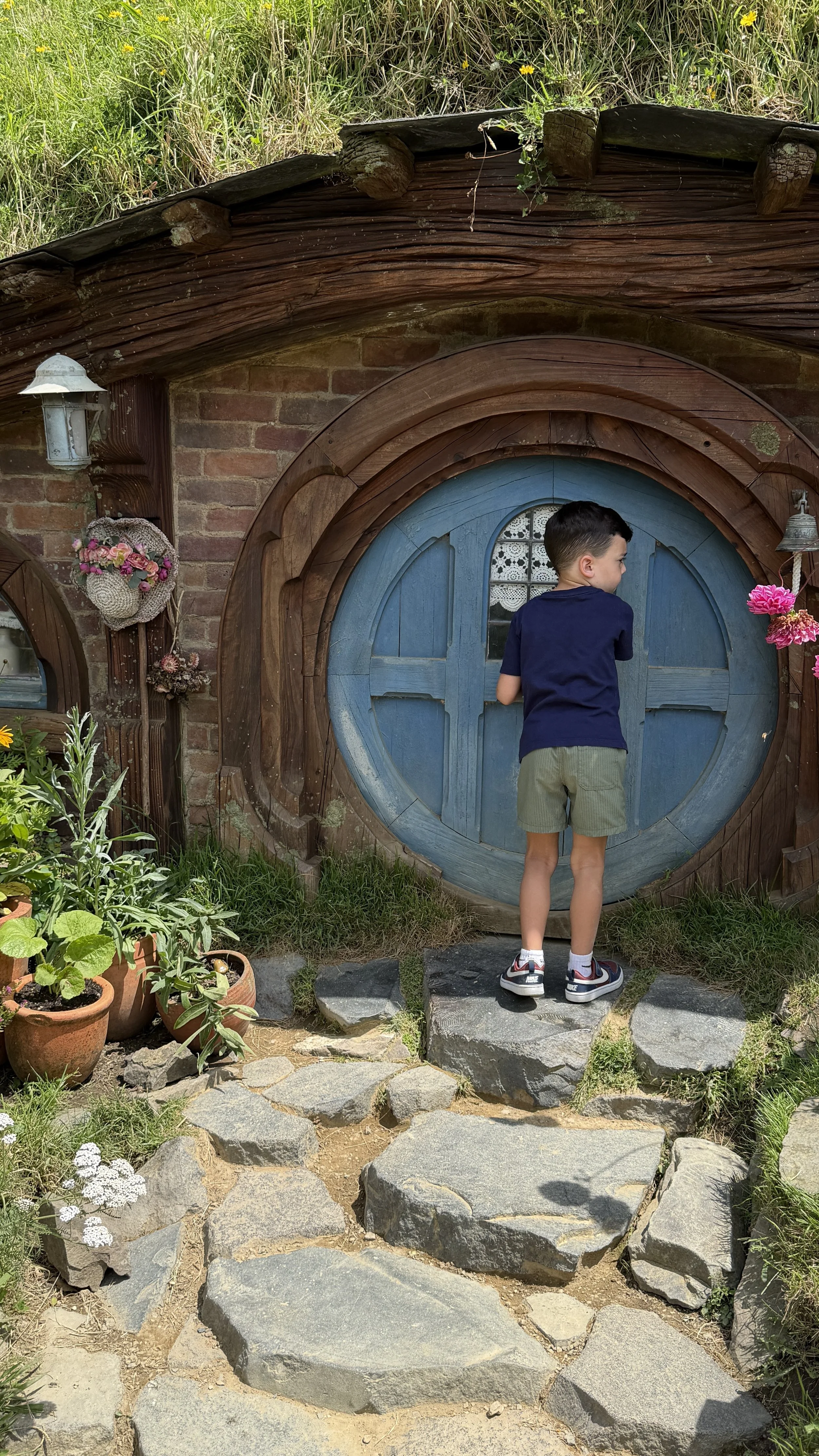 A young boy with short dark hair wearing a navy t-shirt, olive green shorts, and sneakers stands on a stone pathway in front of a small hobbit-style house with a round blue door, brick walls, and a grassy roof adorned with flowers and plants.