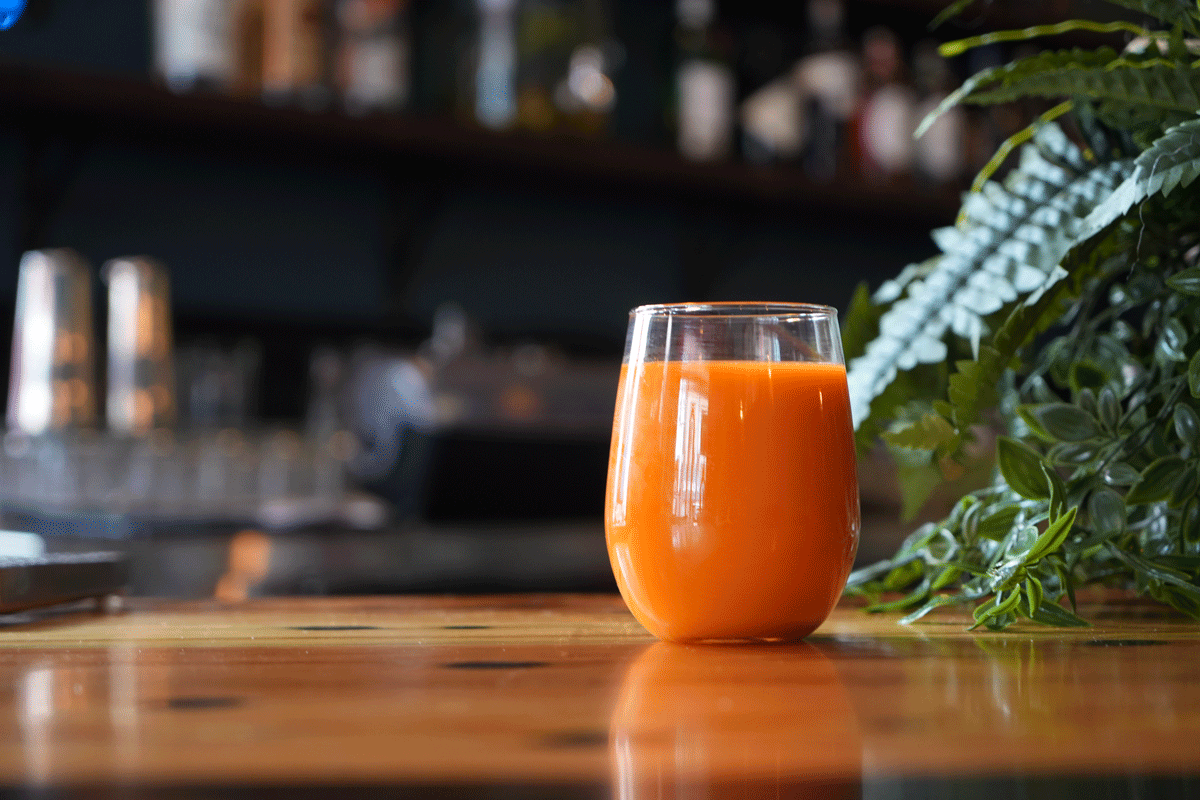 Vibrant orange botanical juice in a stemless glass, set beside greenery on a wood table at Snively’s Lounge.
