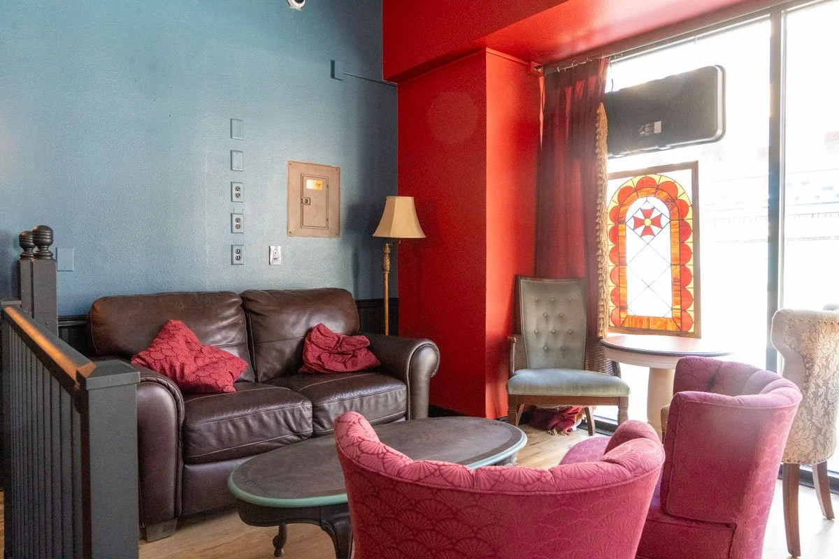 Interior of a cozy lounge with a brown leather sofa, pink pillows, a vintage floor lamp, and a variety of chairs around a dark coffee table. There is a stained-glass window and a large glass door letting in sunlight.