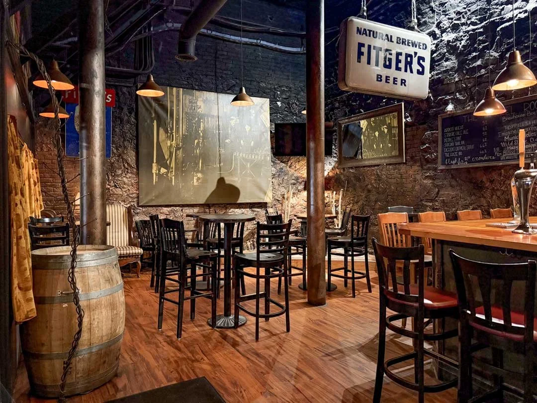 Interior of a rustic bar or pub with wooden floors, high tables and chairs, exposed brick walls, vintage advertising signs, and warm lighting.