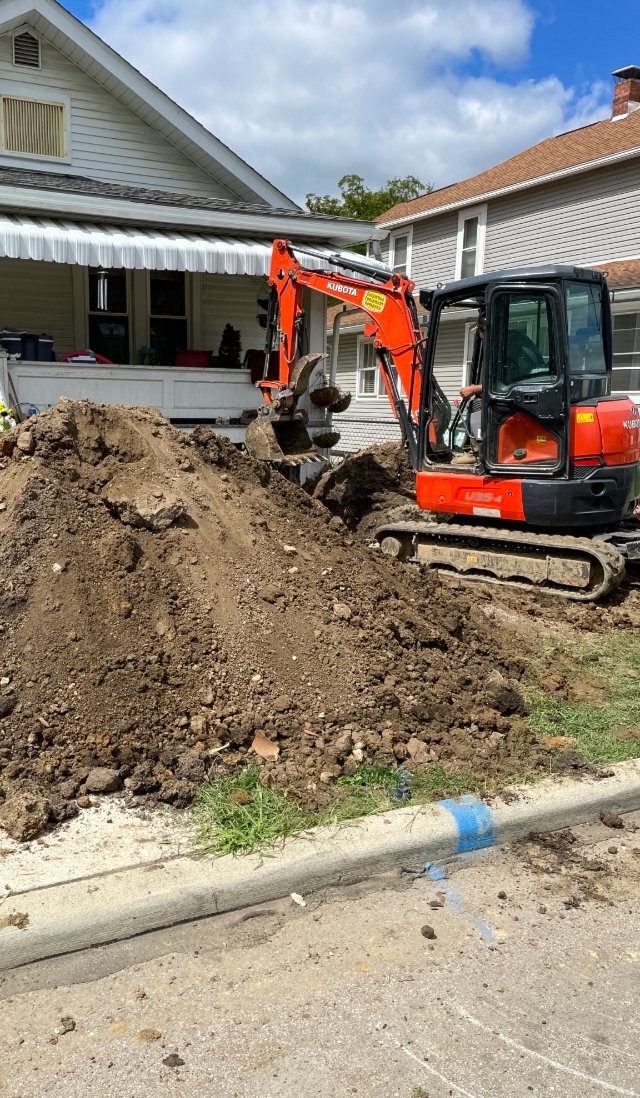 Mini excavator digging dirt pile in front of house on a sunny day.
