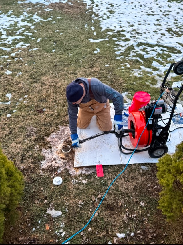 Man wearing work clothes using a drain cleaning machine on a snowy lawn