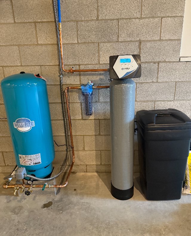 Water filtration and softener system with a blue pressure tank and connected pipes in a cinder block basement.