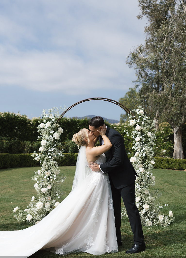 Bride and groom sharing a kiss under a floral arch outdoors during their wedding ceremony.