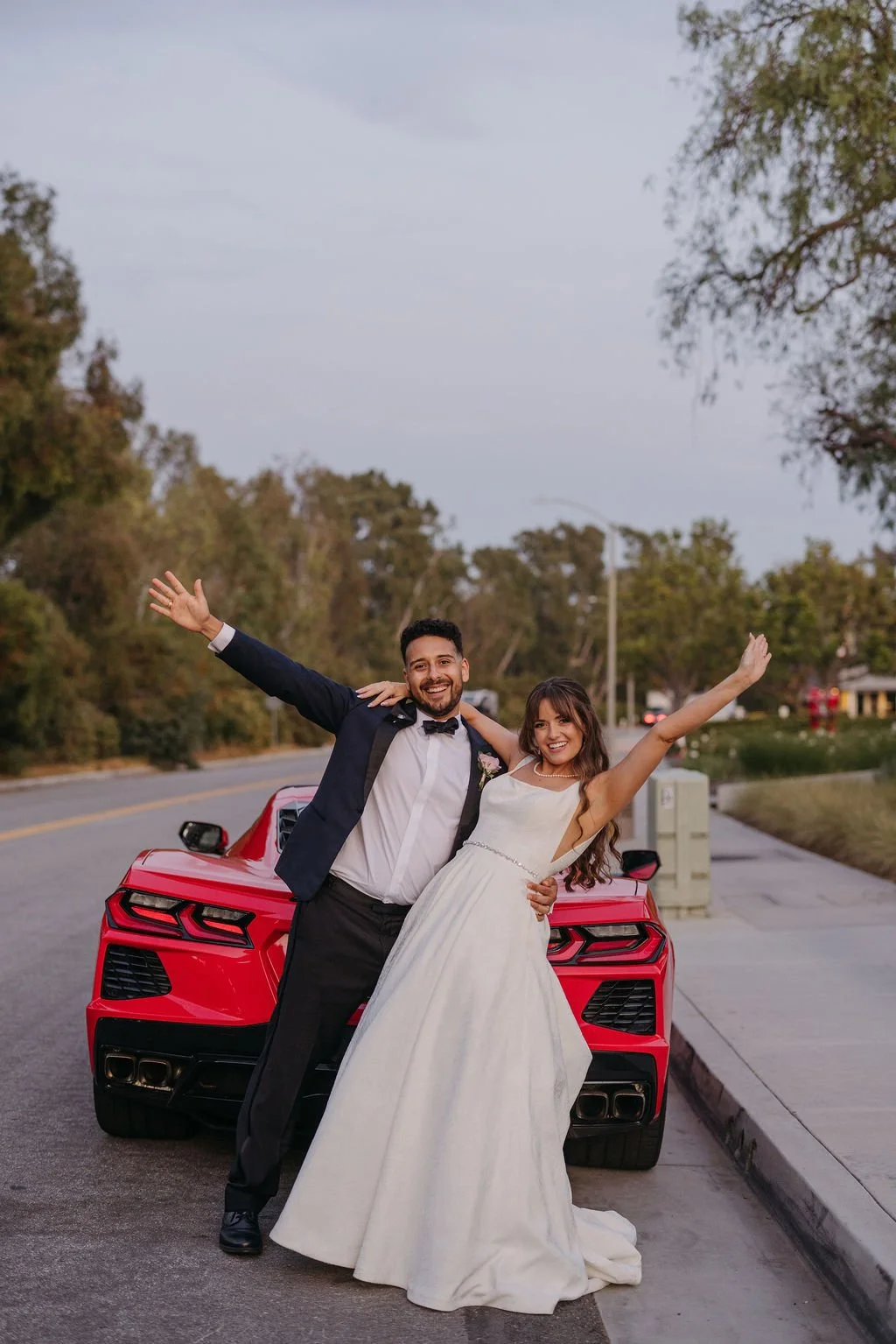 A newlywed couple celebrating in front of a red sports car on a street, with trees and a cloudy sky in the background. The groom is wearing a tuxedo, and the bride is in a white wedding dress. They are smiling and raising their arms.