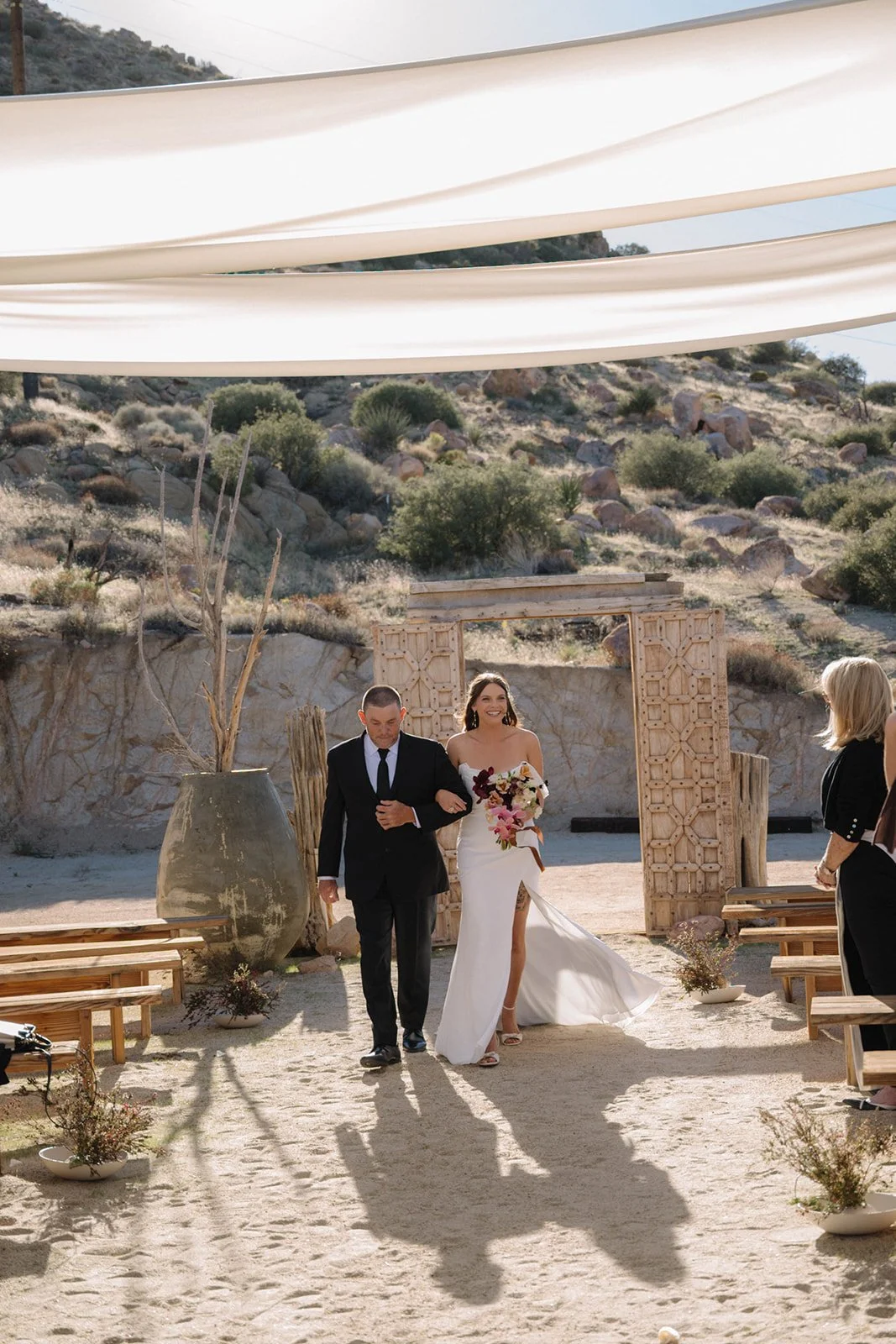A bride walking down the aisle in a desert outdoor wedding with a man, possibly her father, in a black suit. The bride is wearing a white strapless wedding dress with a thigh-high slit and holding a bouquet of flowers. The wedding area has benches, p