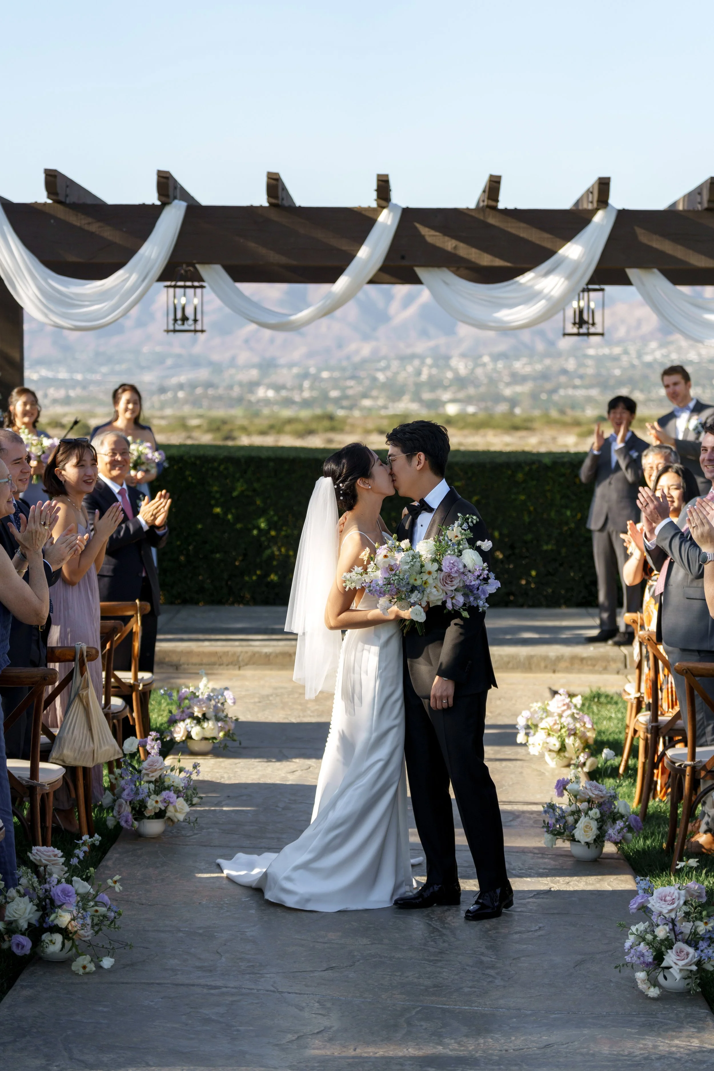 Bride and groom kissing during outdoor wedding ceremony, surrounded by guests clapping and smiling, with decorated floral arrangements and scenic mountain backdrop.