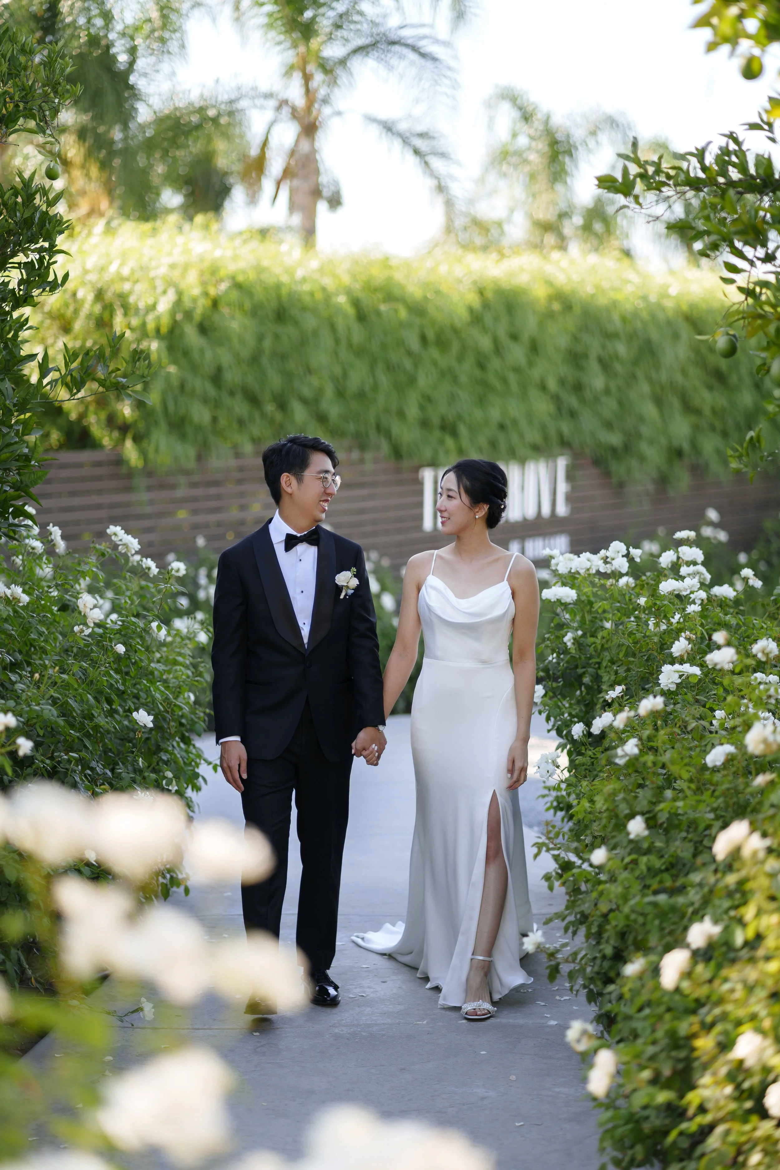 A bride and groom walk hand in hand through a garden pathway, surrounded by white flowers, during their wedding day.