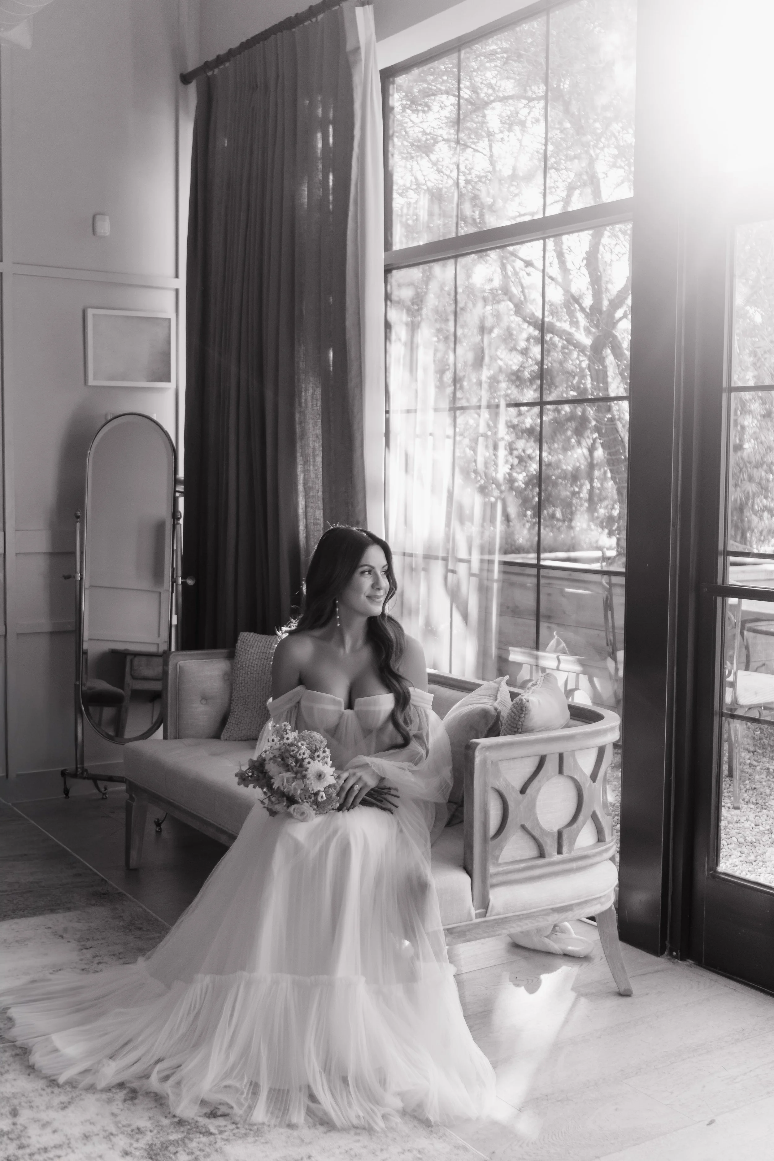 A woman in a wedding dress sitting on a sofa holding a flower bouquet, by a large window with sunlight streaming in.