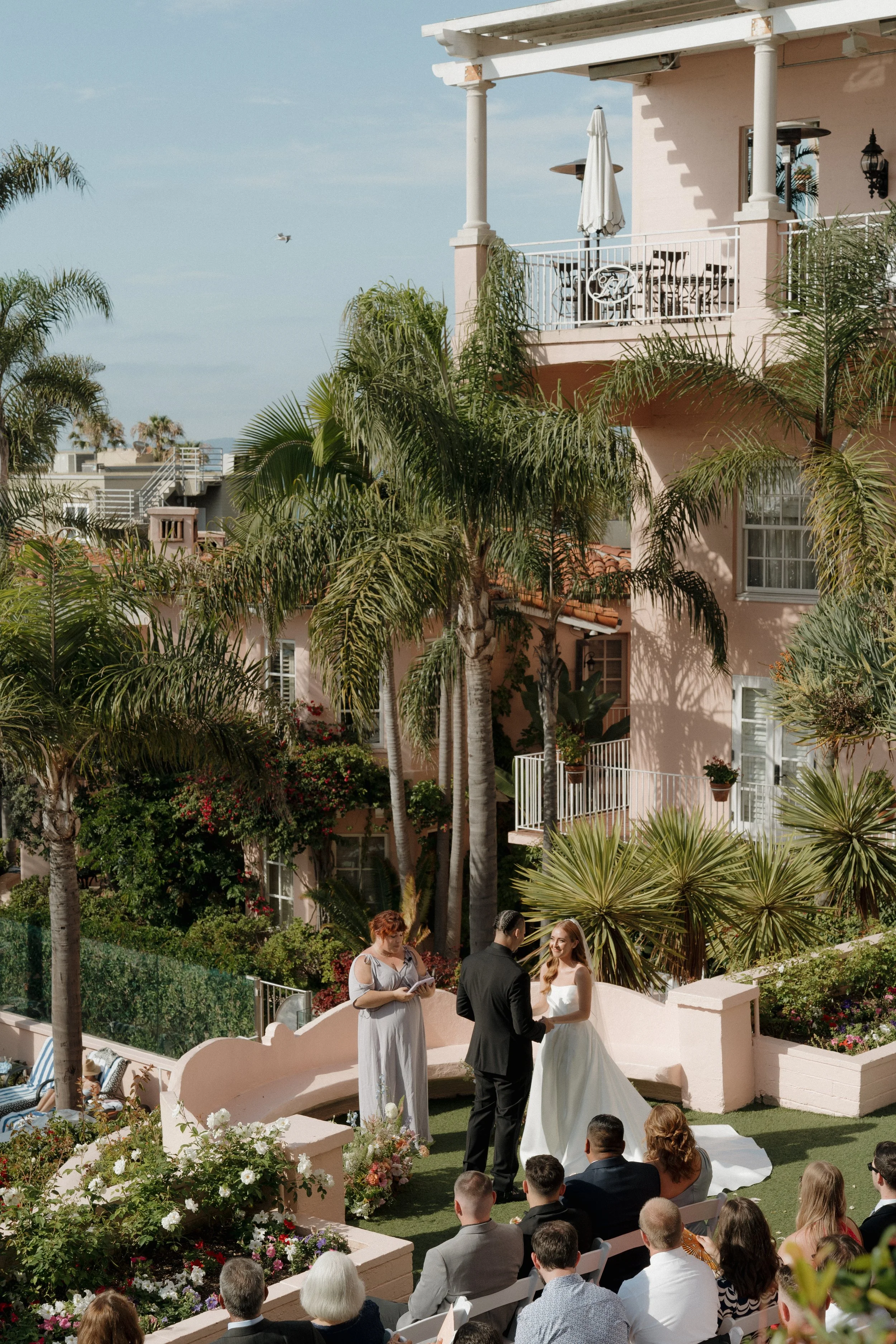 A wedding ceremony taking place outdoors in a garden with pink and green foliage, palm trees, and a pink building with balconies and white railings. The bride in a white dress and the groom in a black suit hold hands in front of an officiant, with gu