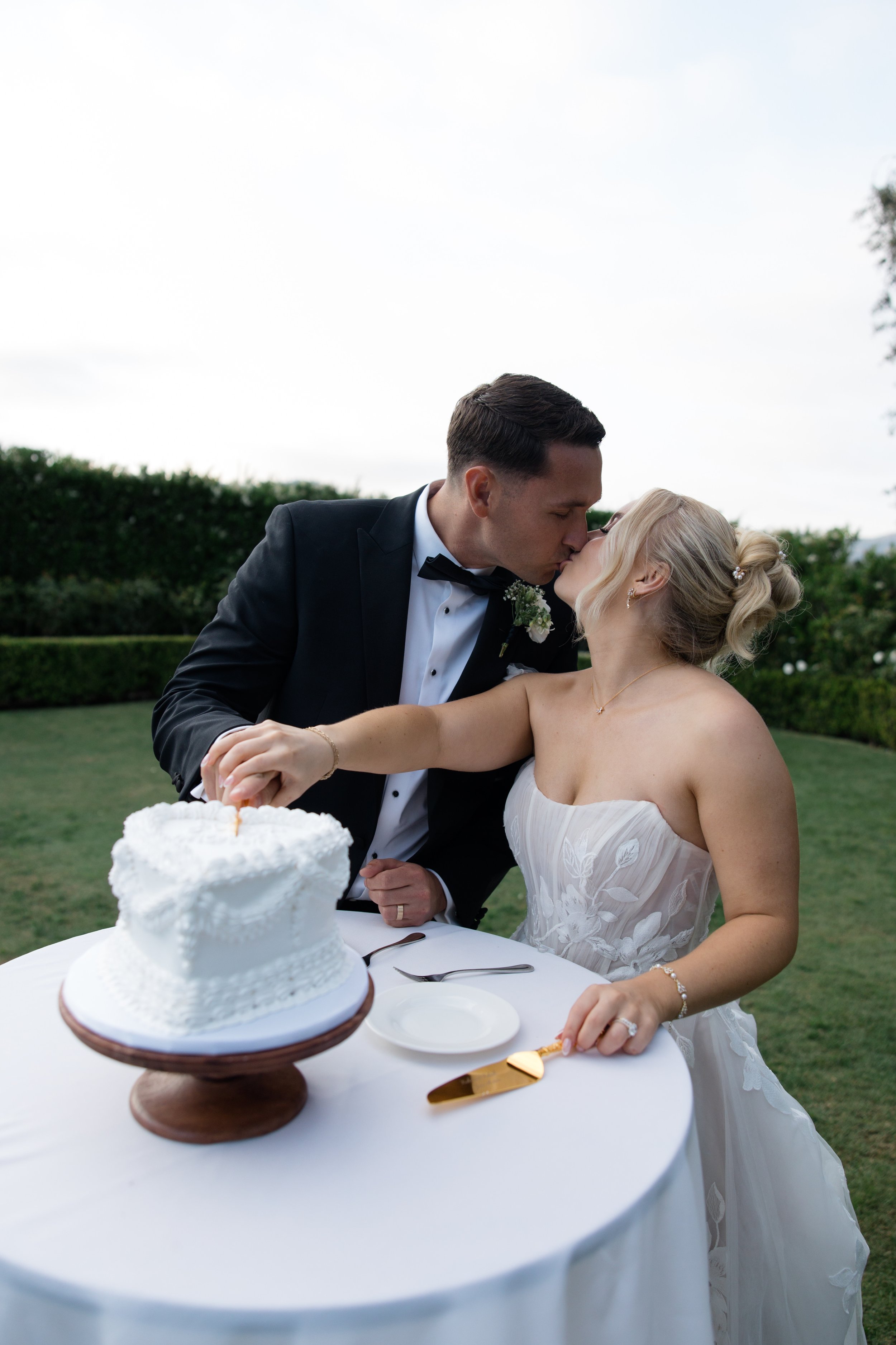 Bride and groom sharing a kiss at their wedding reception, with a wedding cake on the table.