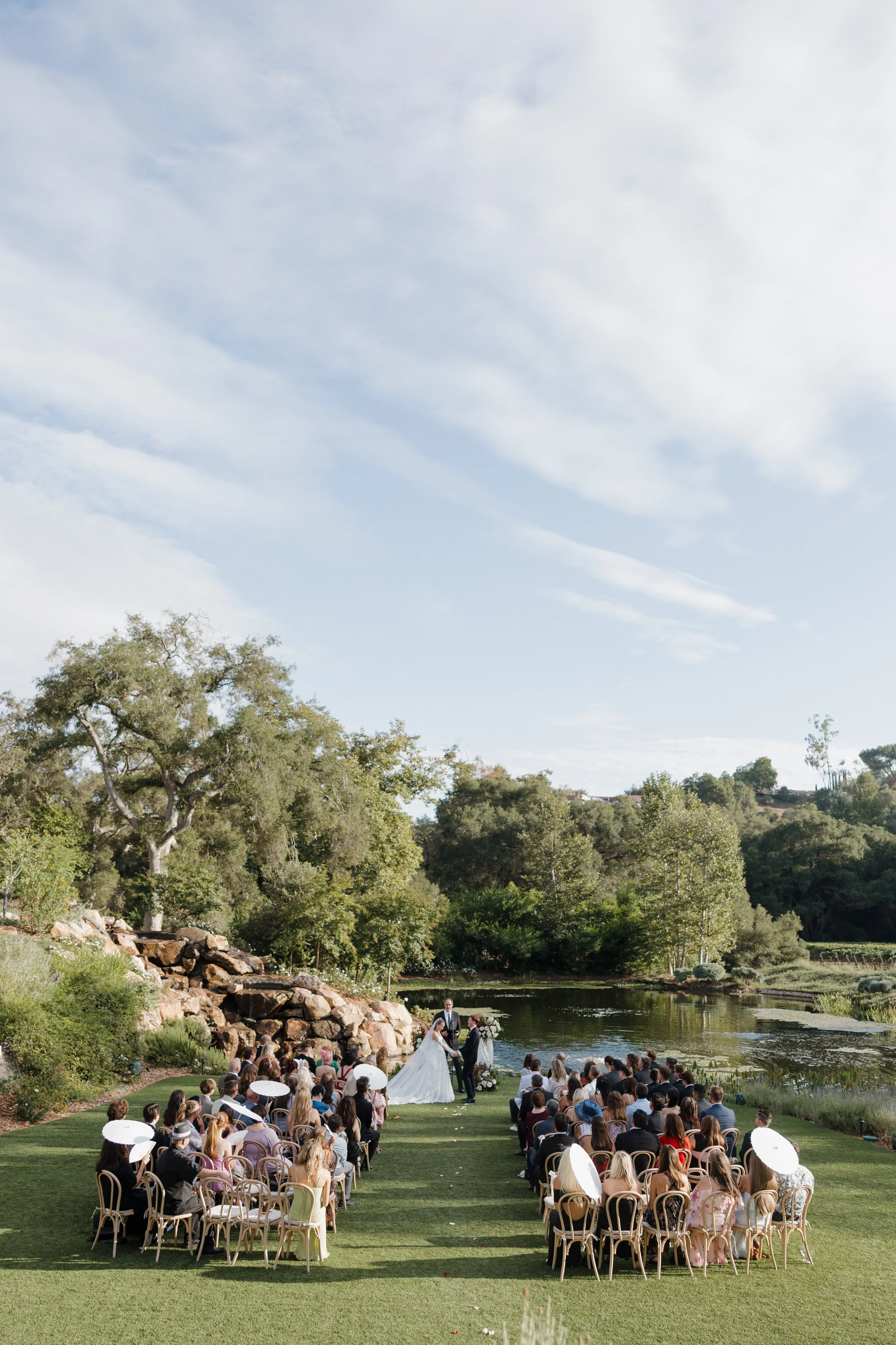 An outdoor wedding ceremony near a lake with guests seated on chairs, the bride and groom holding hands at the altar.