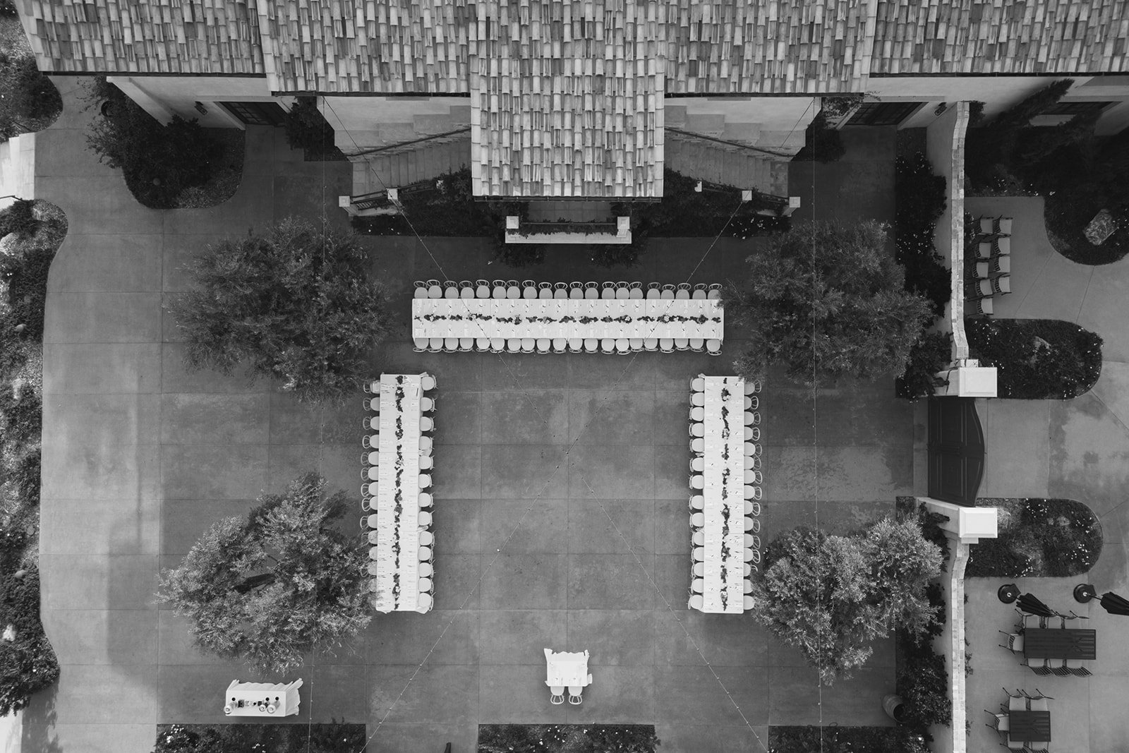 Black and white aerial view of a courtyard with long tables, chairs, trees, and surrounding buildings, set up for an outdoor event or gathering.