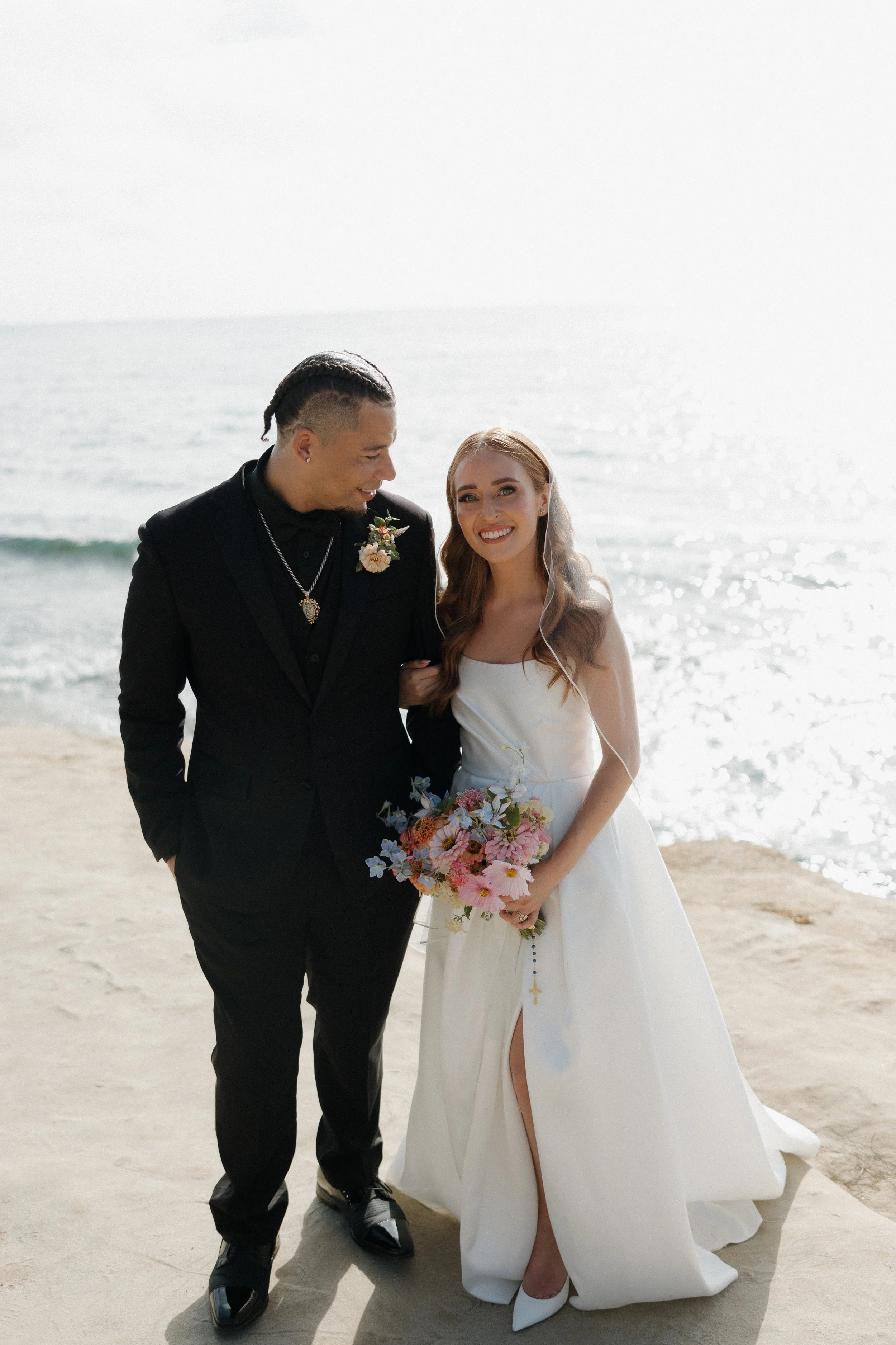 A bride and groom stand on a beach during their wedding, smiling at each other. The bride wears a white gown and holds a colorful bouquet, while the groom wears a black suit and has a boutonniere.