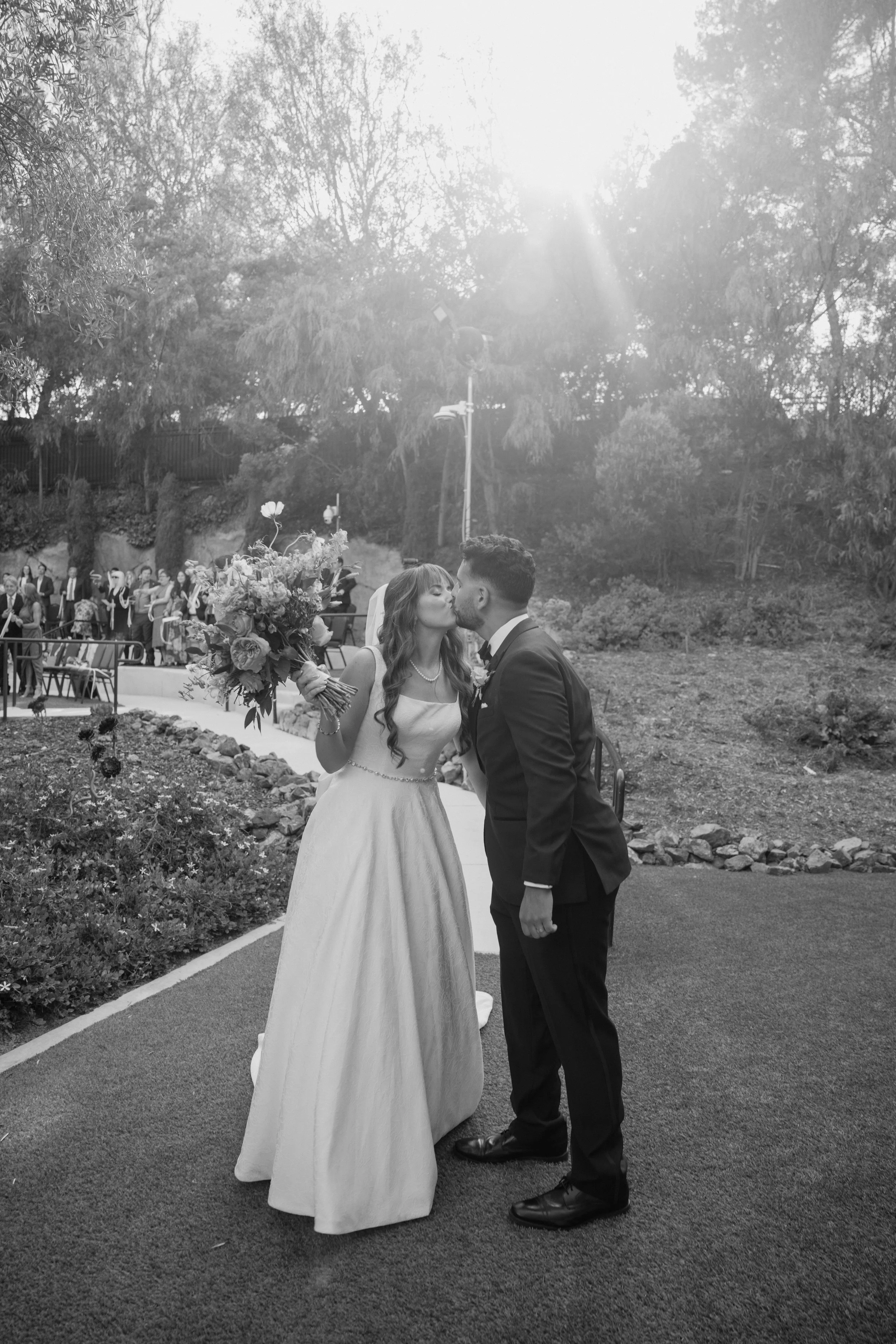 Bride and groom share a kiss at their outdoor wedding, bride holding a bouquet, surrounded by wedding guests and lush greenery.