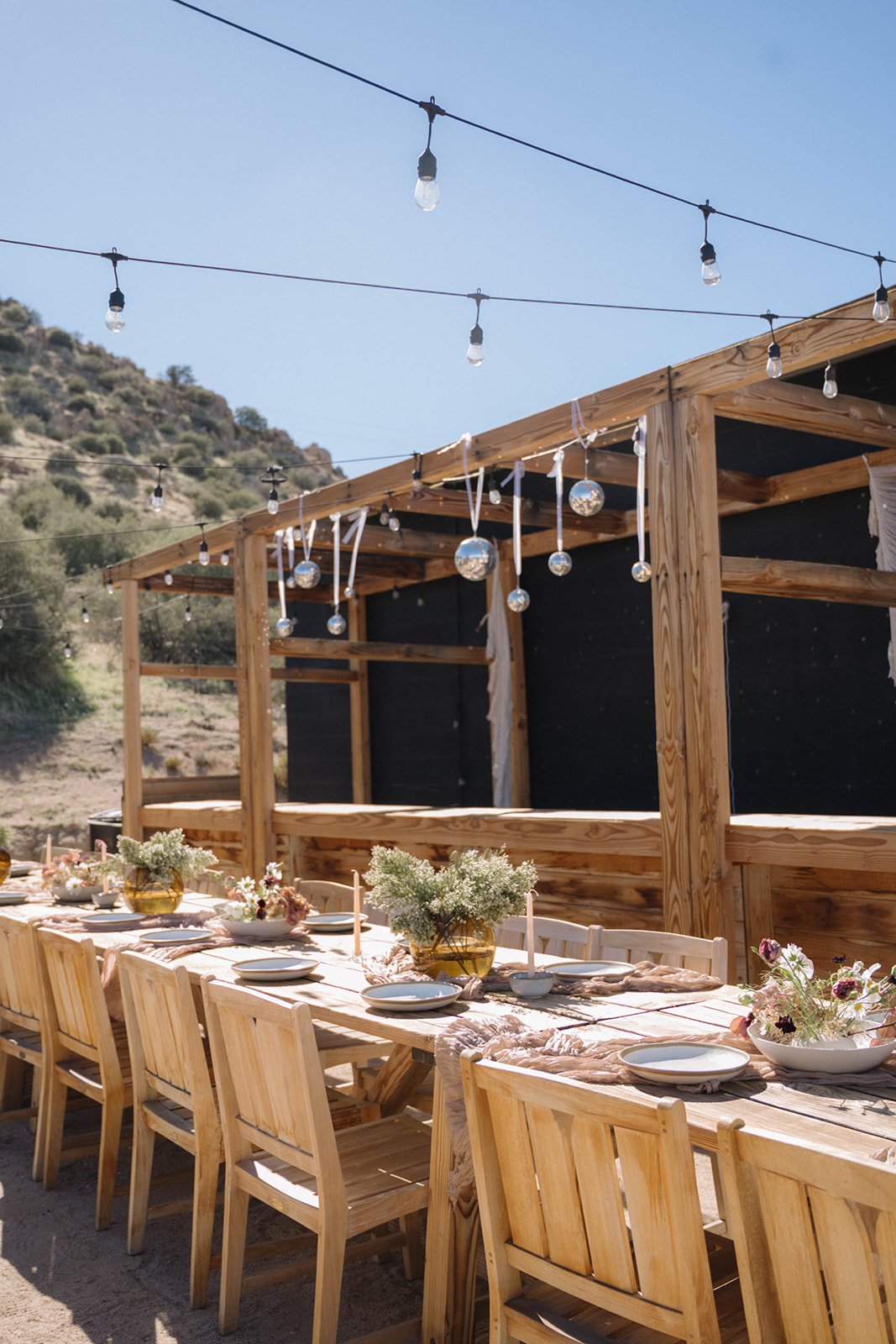 Outdoor wooden dining table decorated with flowers and plates, set under string lights with a clear sky and mountainous landscape in the background.