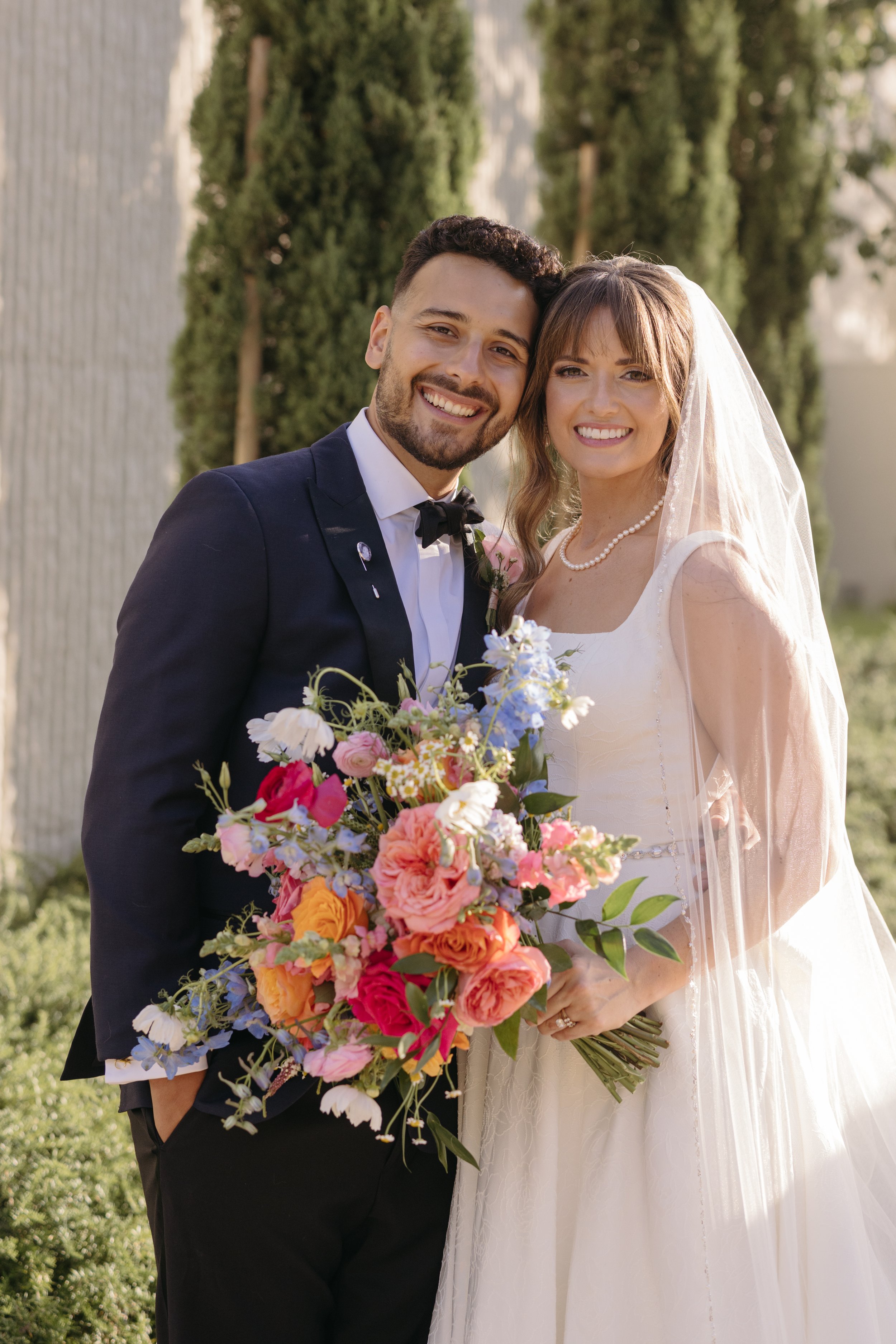 A happy bride and groom smiling outdoors on their wedding day, with the bride holding a large colorful bouquet and wearing a wedding dress, veil, and pearl necklace, while the groom is dressed in a dark suit with a bow tie.