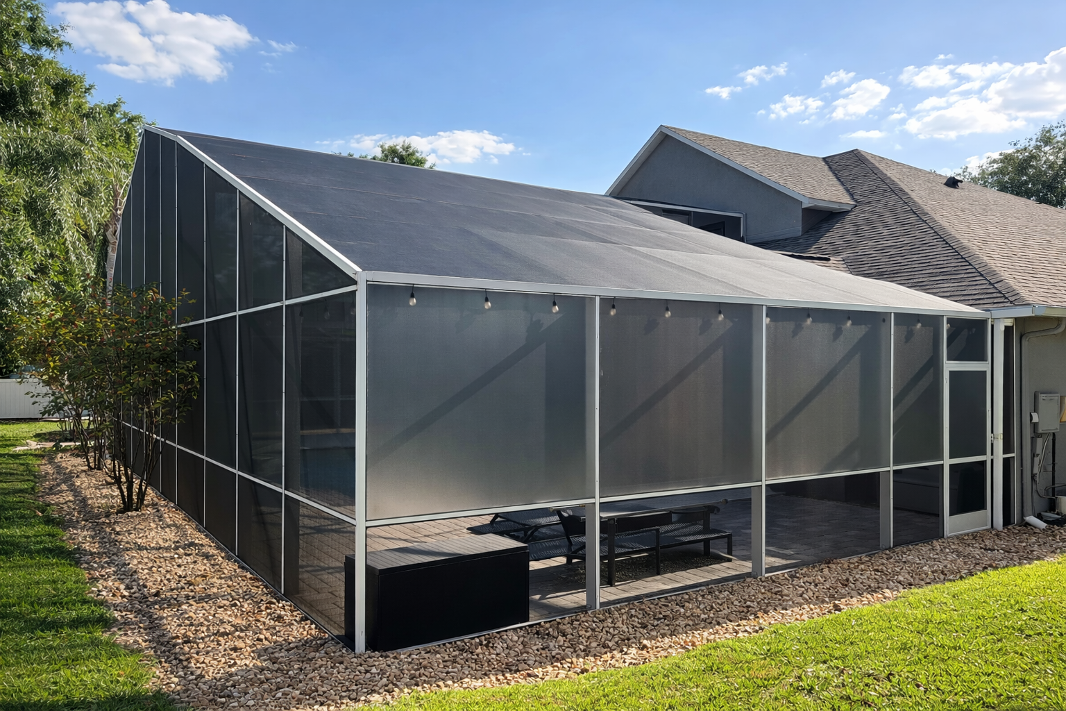 A screened-in patio enclosure attached to a house, with outdoor furniture and string lights, surrounded by a grassy lawn, trees, and a blue sky.