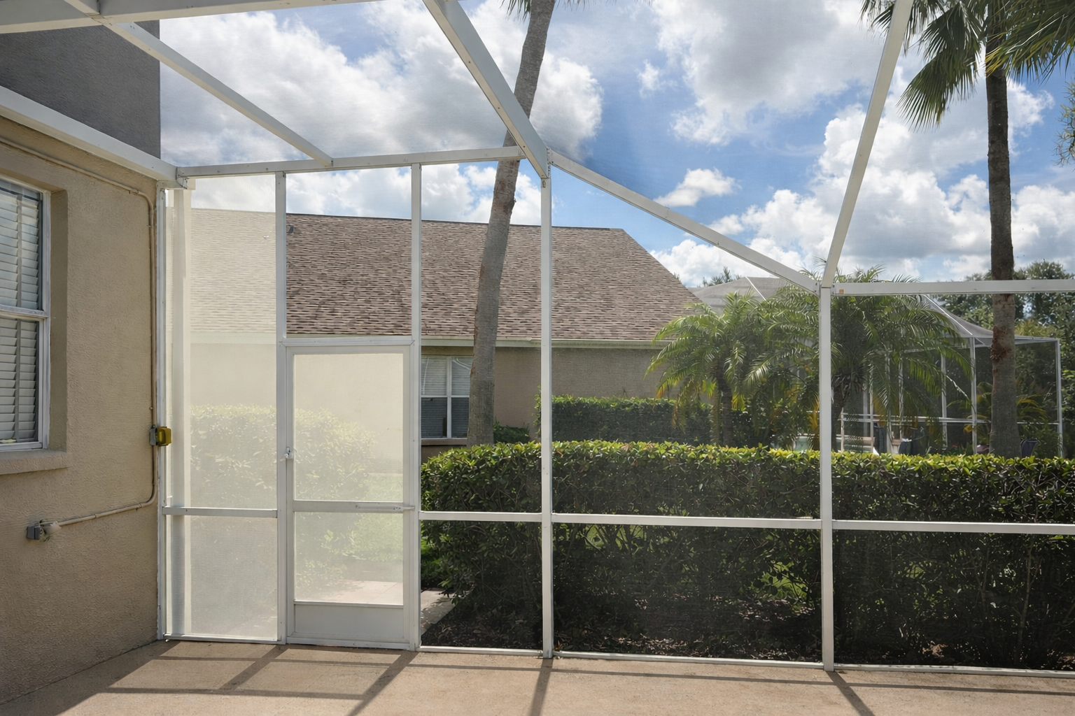 View of an enclosed backyard patio with a screened door, lush green bushes, palm trees, and a rooftop visible in the background under a partly cloudy sky.