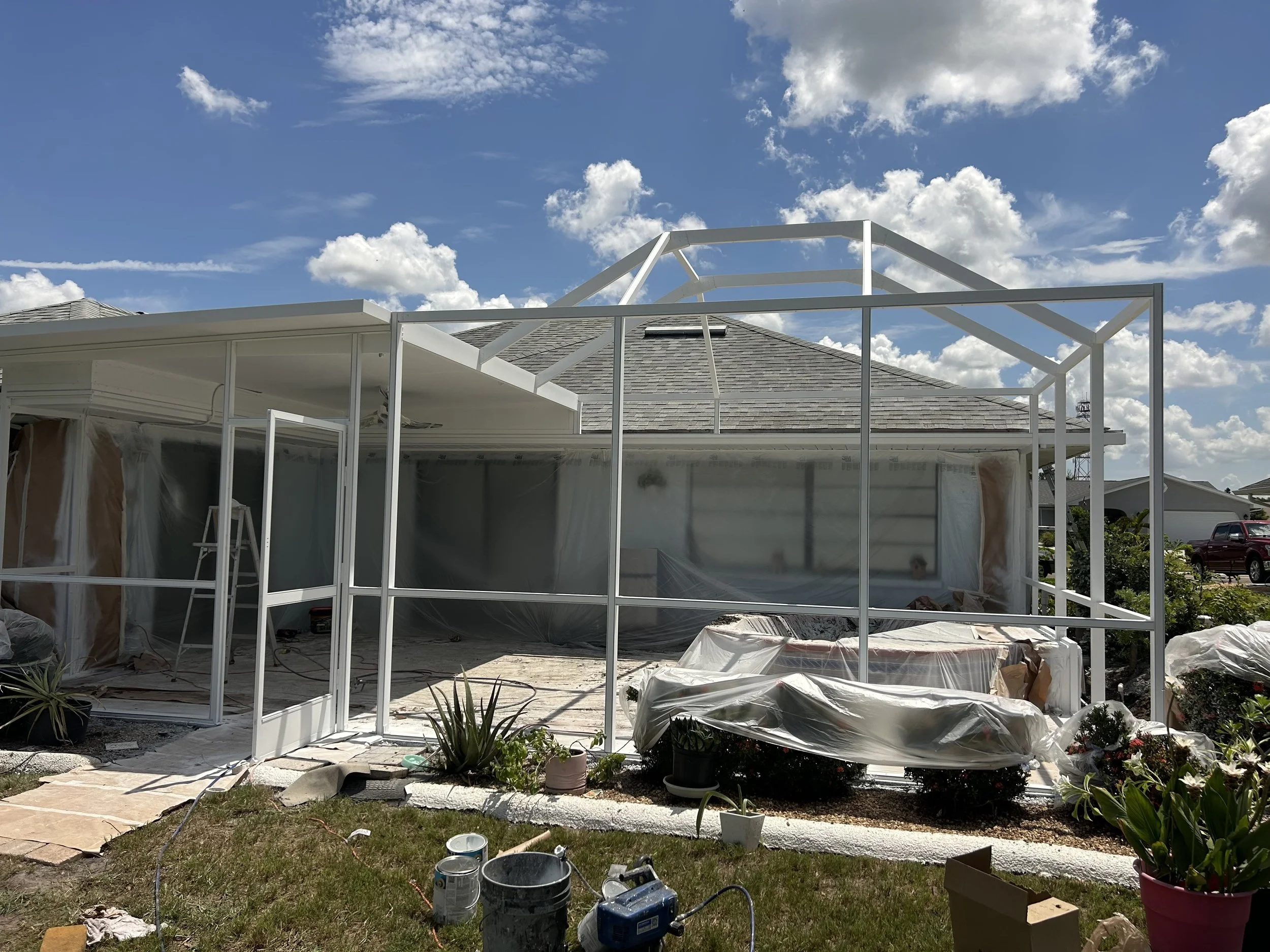 Construction of a screened porch or patio extension on a house with a gray shingle roof, white framing, and some patio furniture covered with plastic, with a partly cloudy sky above.