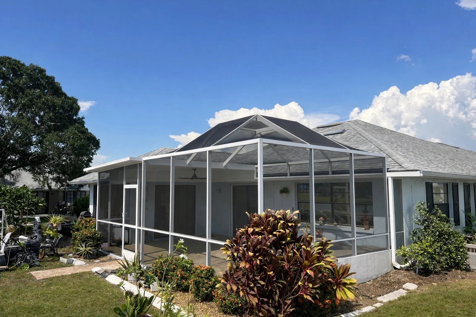 A house with a screened porch and a garden with various plants and bushes under a blue sky with clouds.