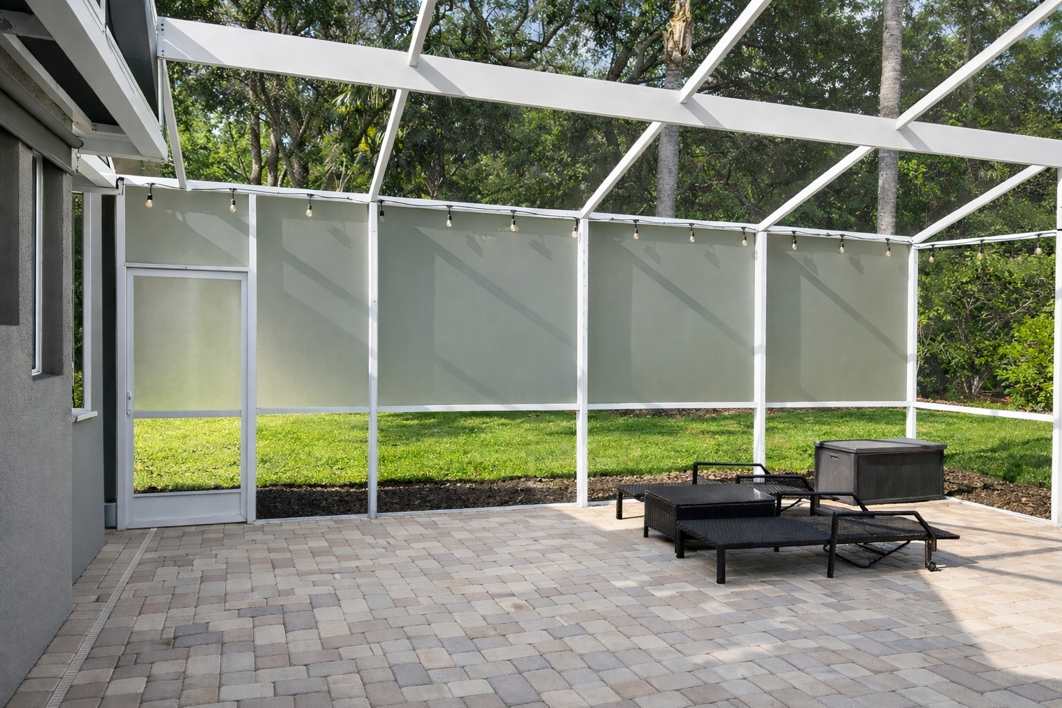 Enclosed backyard patio with paver stones, outdoor furniture, and string lights, surrounded by trees and greenery.