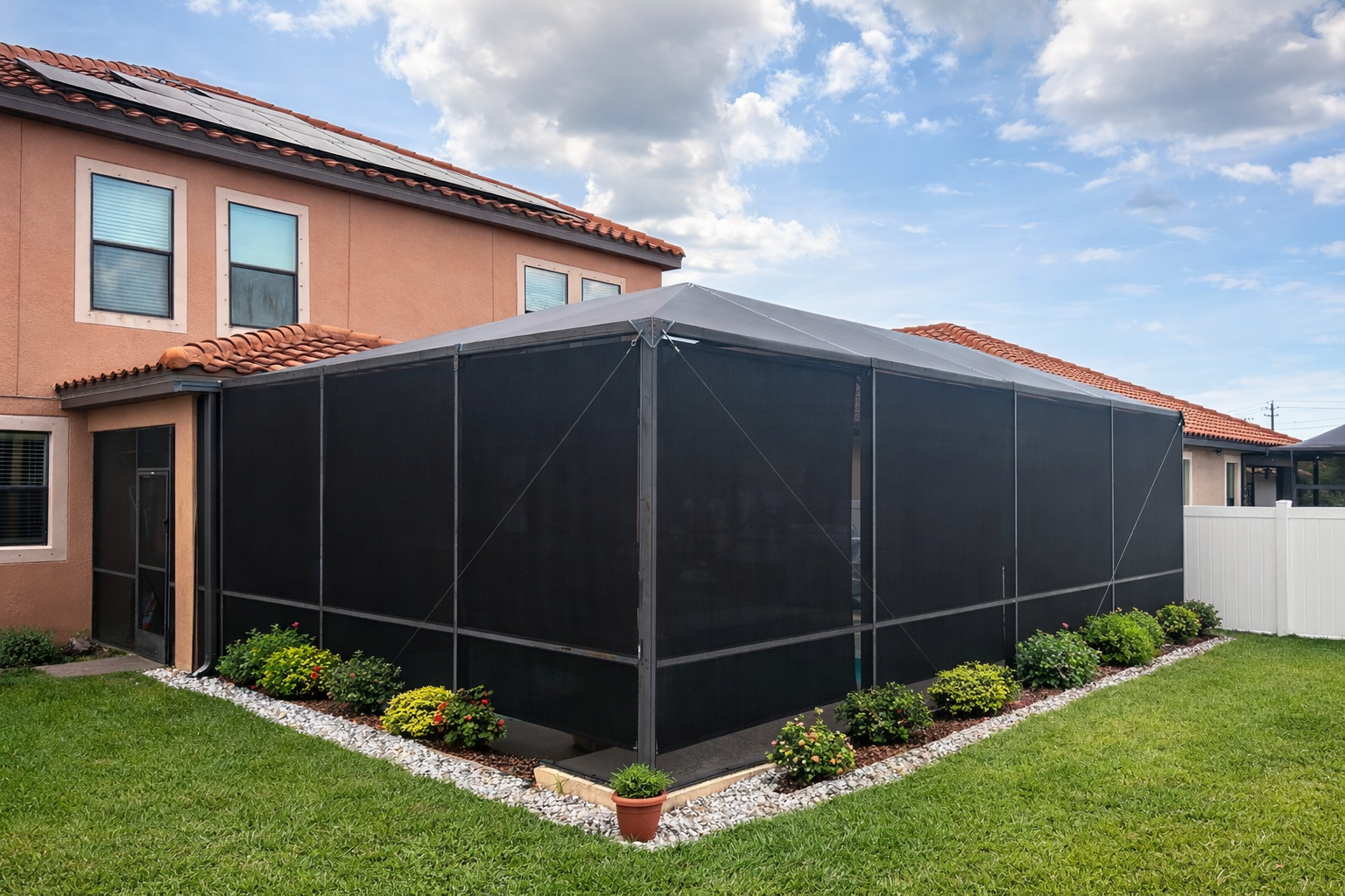 Backyard with a screened-in porch attached to a two-story house with orange stucco walls and a red tiled roof. The porch has black screening and is surrounded by well-maintained green grass, small bushes, and decorative stones.