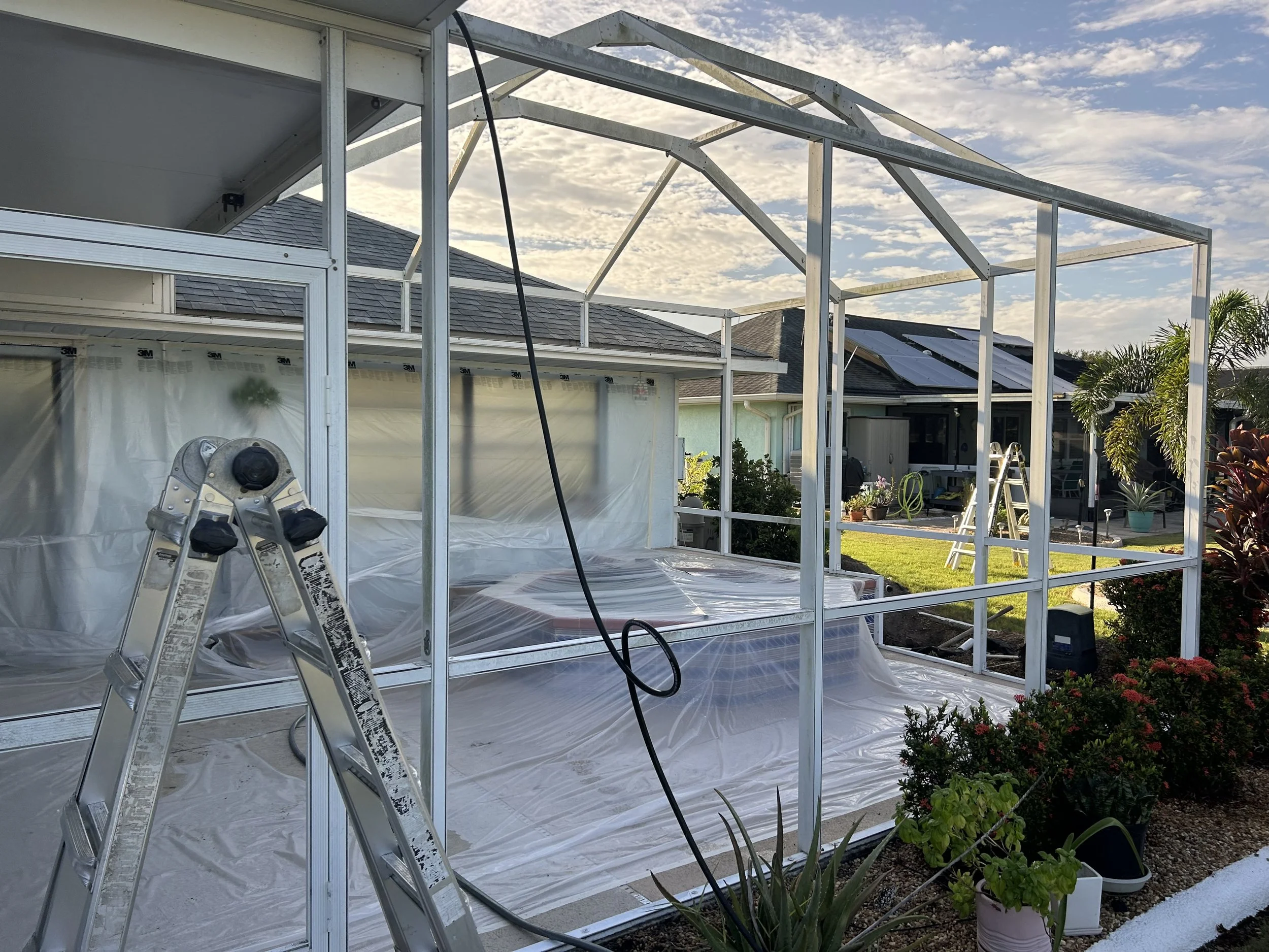A metal frame structure is being built for a backyard patio or enclosure with a ladder and tools visible. Plastic sheeting covers the ground inside the frame, and there are potted plants along the edge of the construction area. Neighboring houses with solar panels on the roofs are visible in the background under a partly cloudy sky.