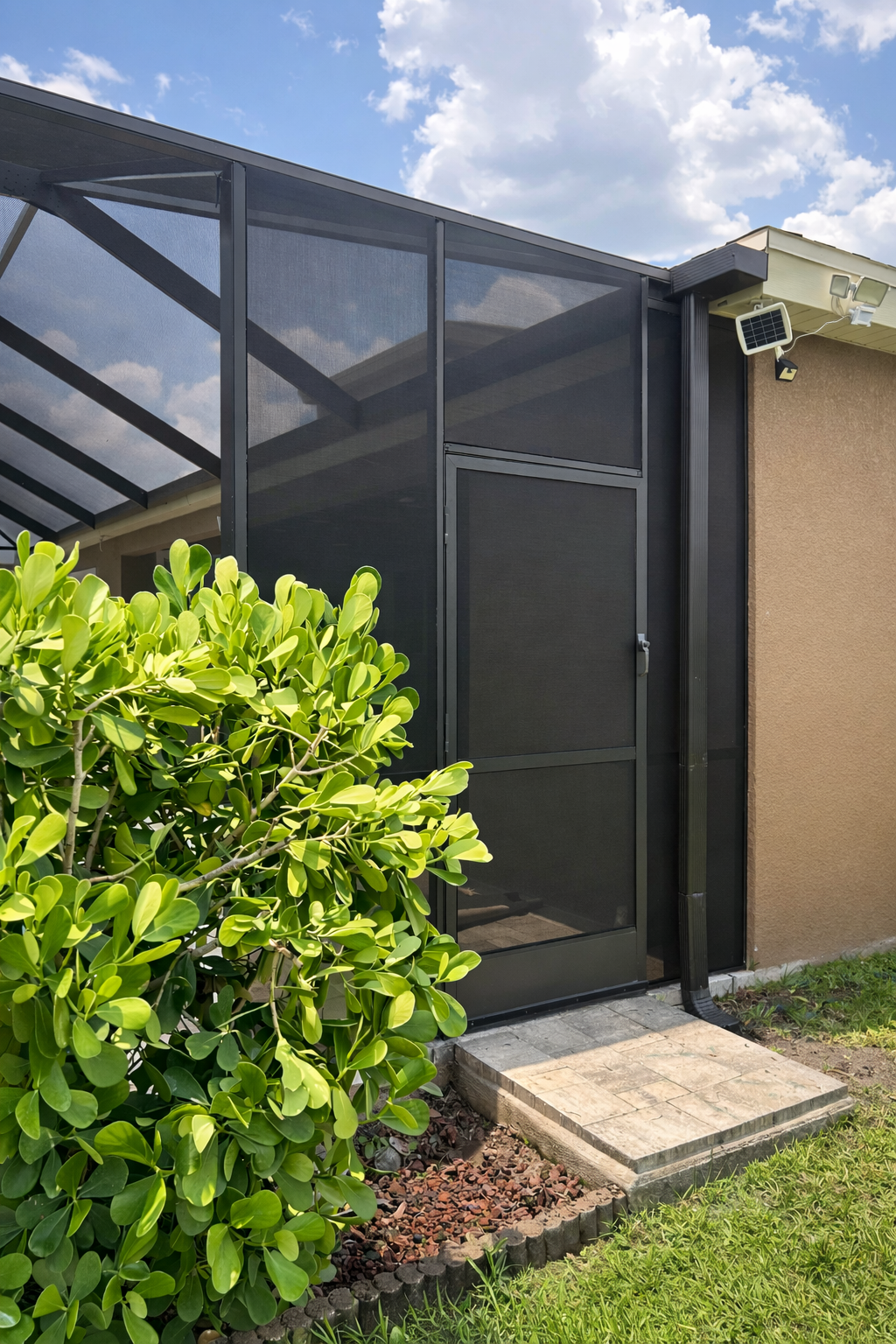 A screened outdoor patio area attached to a house, with a black metal door, a small solar-powered light, and security camera, surrounded by greenery and a partly cloudy sky.