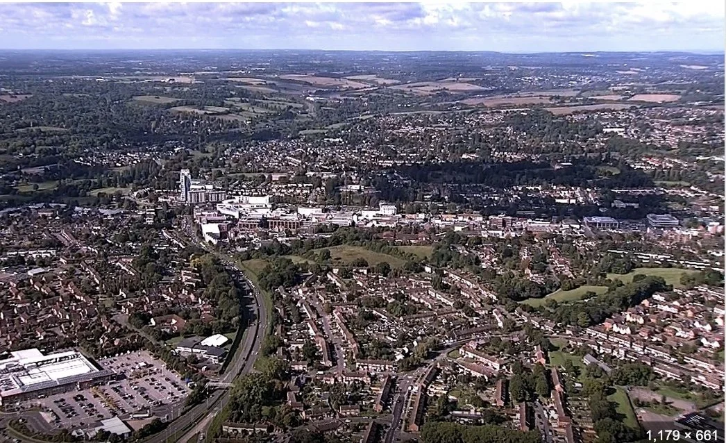 Aerial view of hertfordshire with a mix of residential neighborhoods, commercial buildings, and green spaces under a partly cloudy sky.