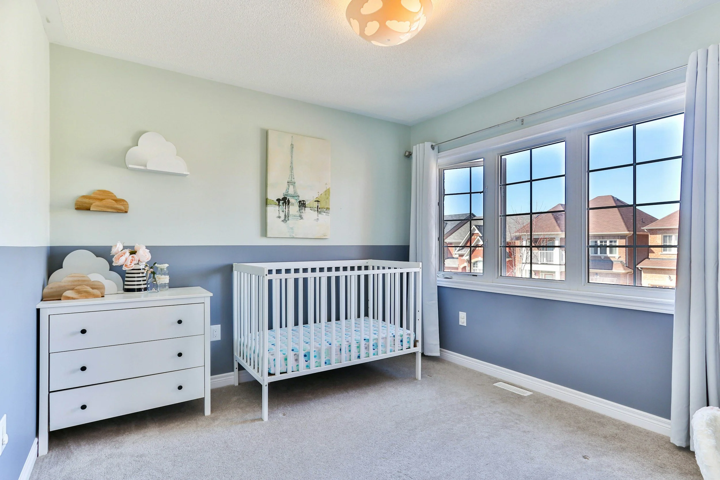 Bright blue baby nursery with a crib, dresser, and large windows