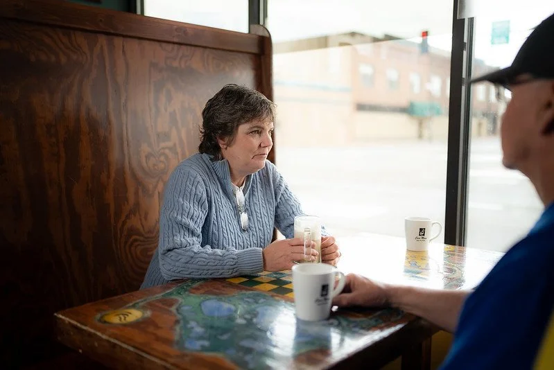 A woman sitting at a table inside a cafe, holding a glass of water, with a man partially visible holding a coffee mug across from her.