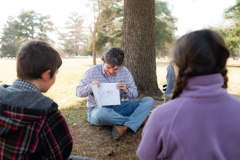 A person with short gray hair sitting under a tree, holding an open sketchbook and demonstrating drawings to three children outdoors on a sunny day.
