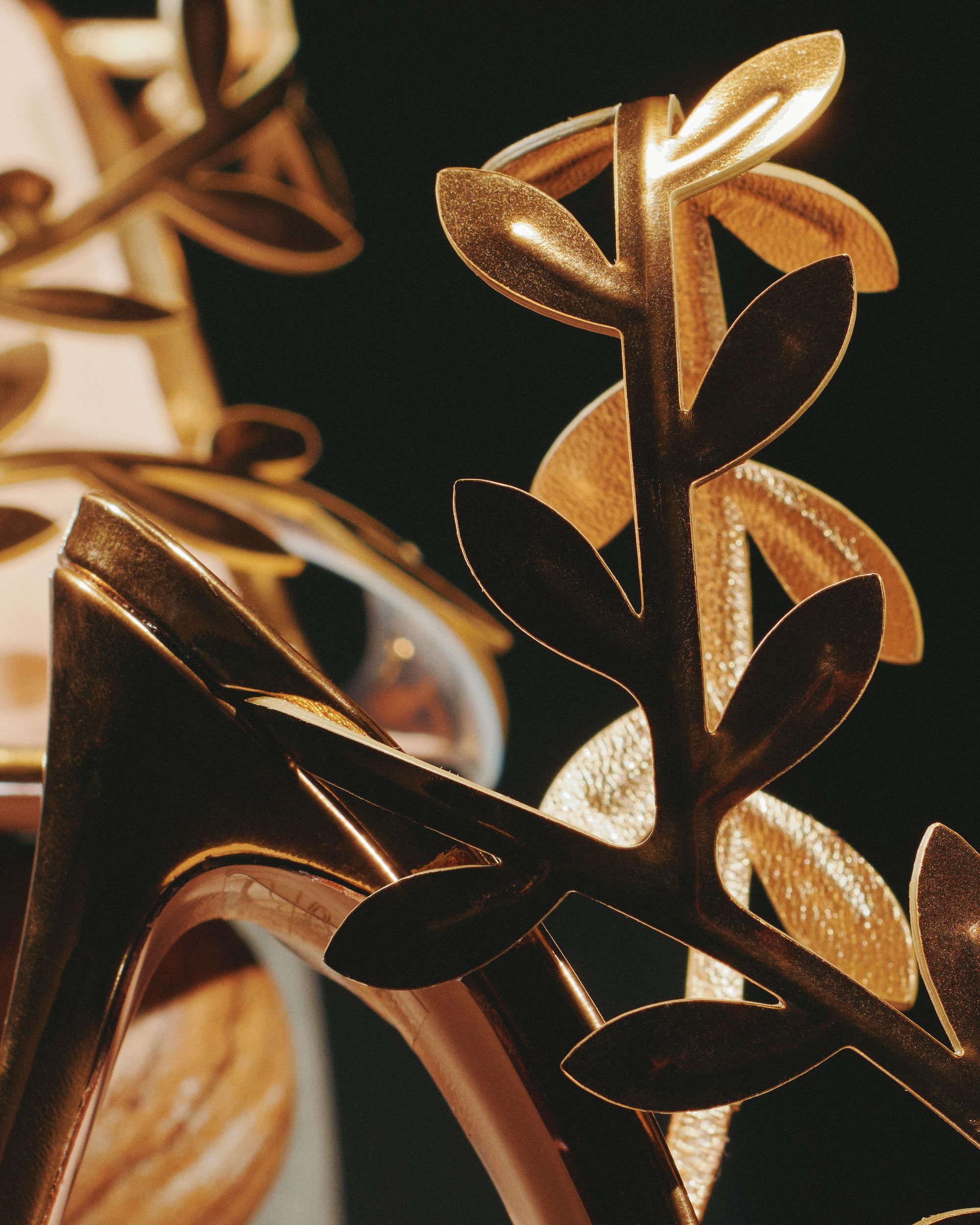 Close-up of gold and metallic decorative accessories, including a gold leaf-shaped ornament and a pair of gold high-heeled shoes, against a dark background.