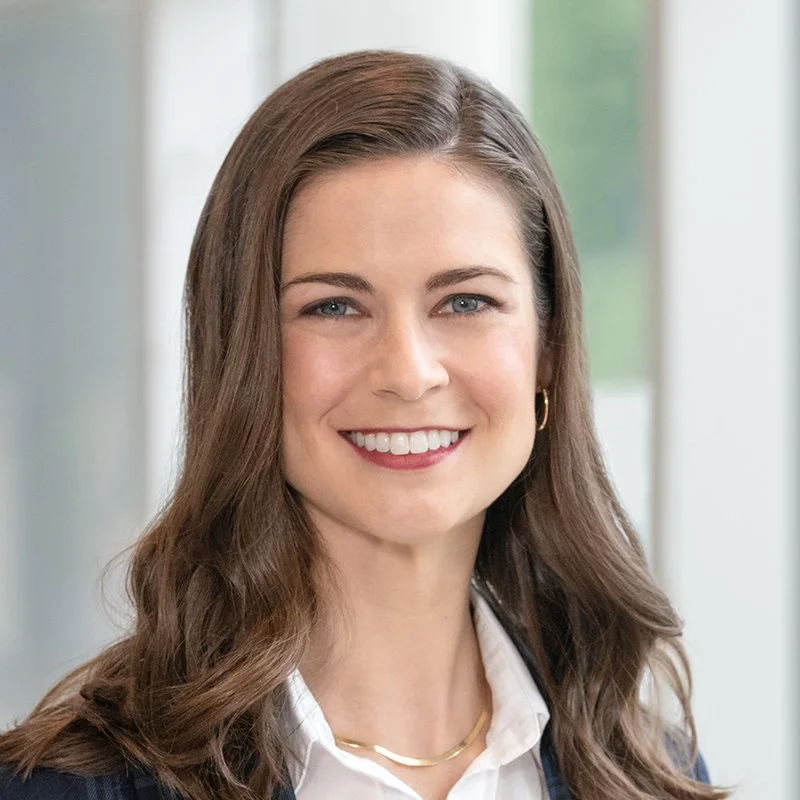A woman with long brown hair and blue eyes smiling in a professional setting, wearing a white shirt, navy blazer, and gold jewelry.