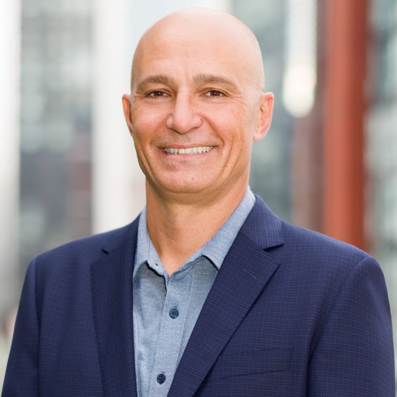 Headshot of a bald, smiling man in a blue suit and light blue shirt, standing outdoors with a blurred background of tall glass buildings.