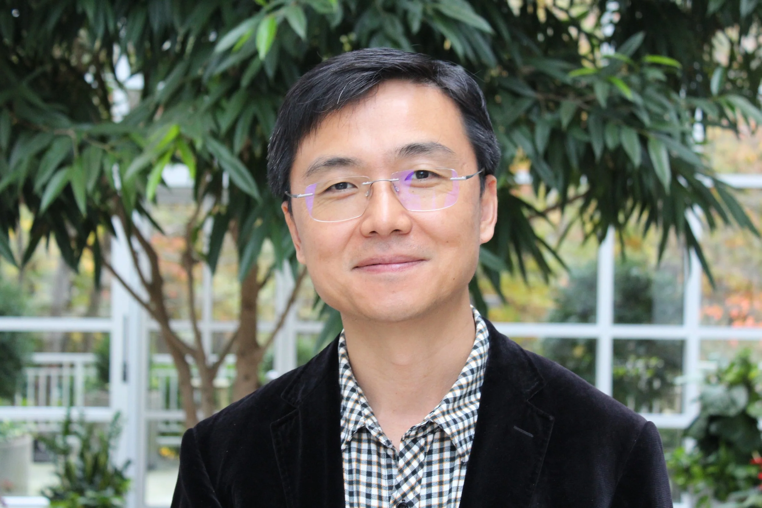 A portrait of a man with black hair, glasses, and a slight smile, wearing a black suit jacket and a white shirt, standing against a light background.